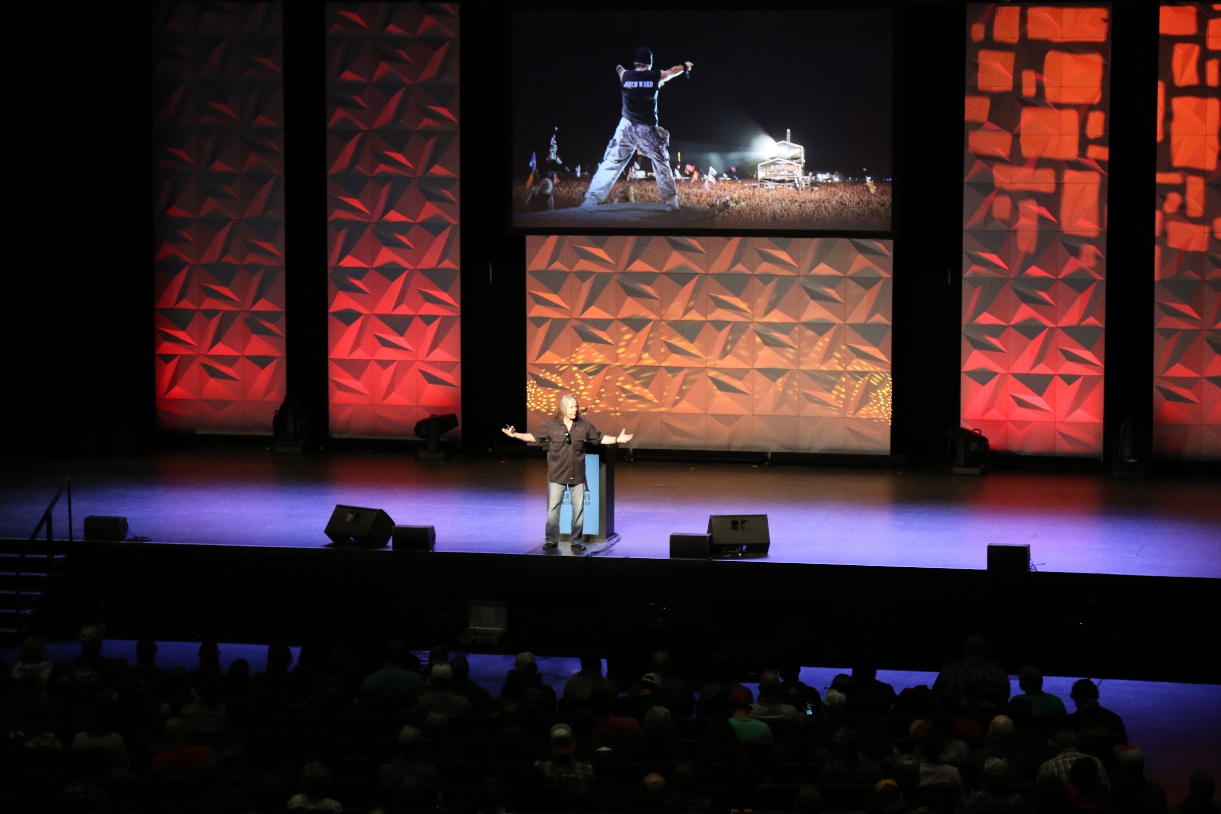 A speaker with outstretched arms on a lit stage, with a large screen above showing a performer on a stage at night.