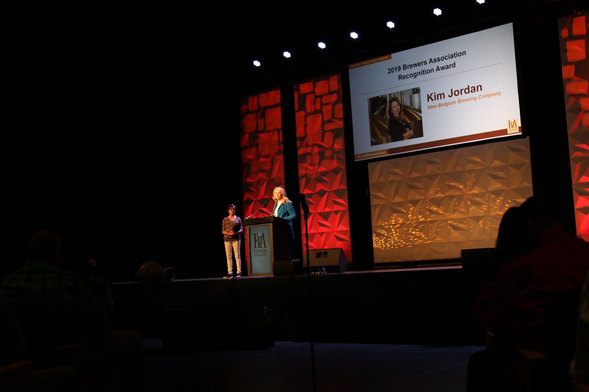 Two people at a podium beside a screen reading 2019 Brewers Association Recognition Award, Kim Jordan, New Belgium Brewing Company.