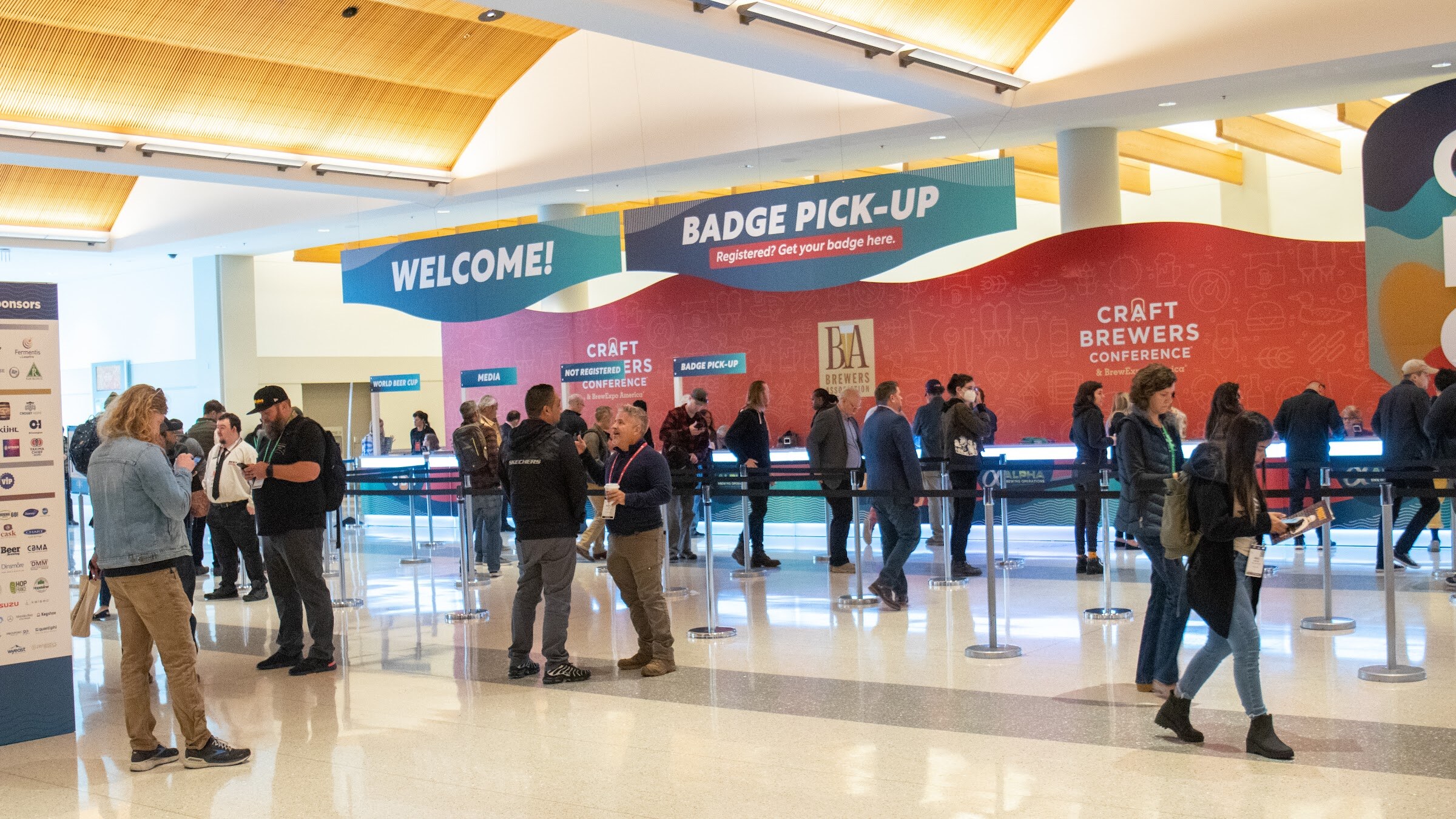 Attendees queue at a long registration counter under a red and white welcome and badge pick-up banner