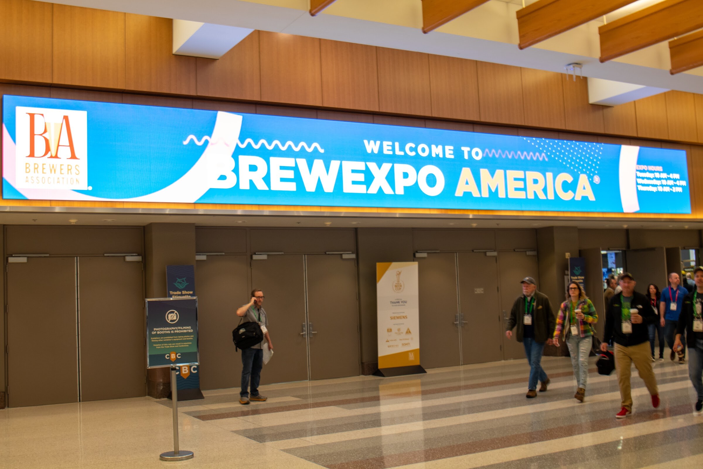 A wide LED screen reading Welcome to BrewExpo America hangs over the entrance doors while attendees walk past