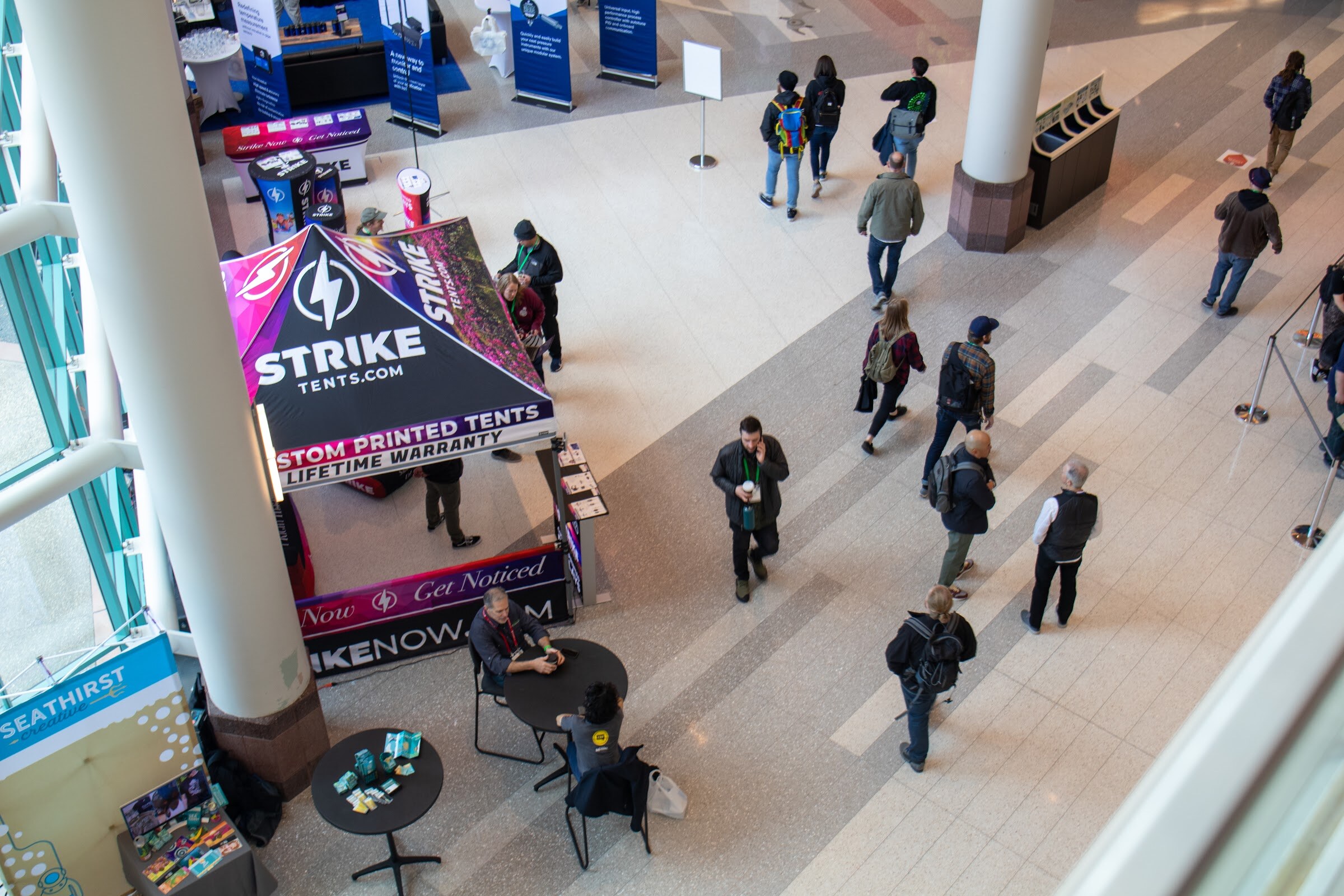 Looking down from above, attendees walk past a Strike Tents display and small vendor tables on the convention center lobby floor