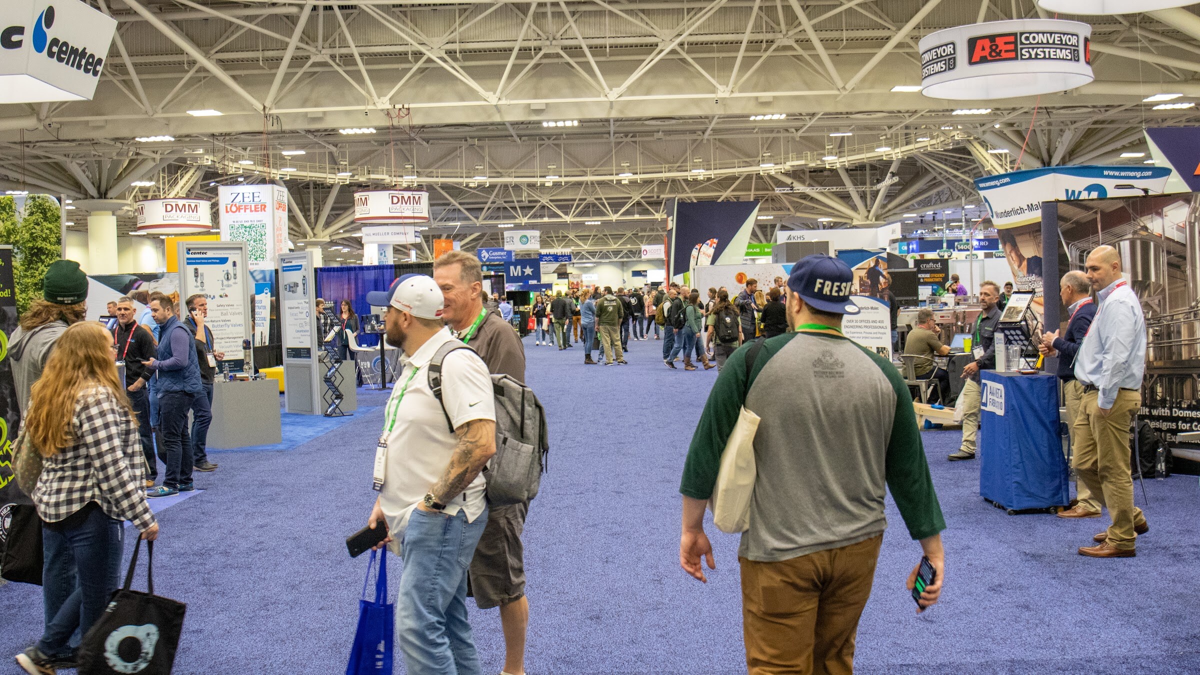 Attendees walk down an aisle of blue carpet between supplier booths under the convention center rafters