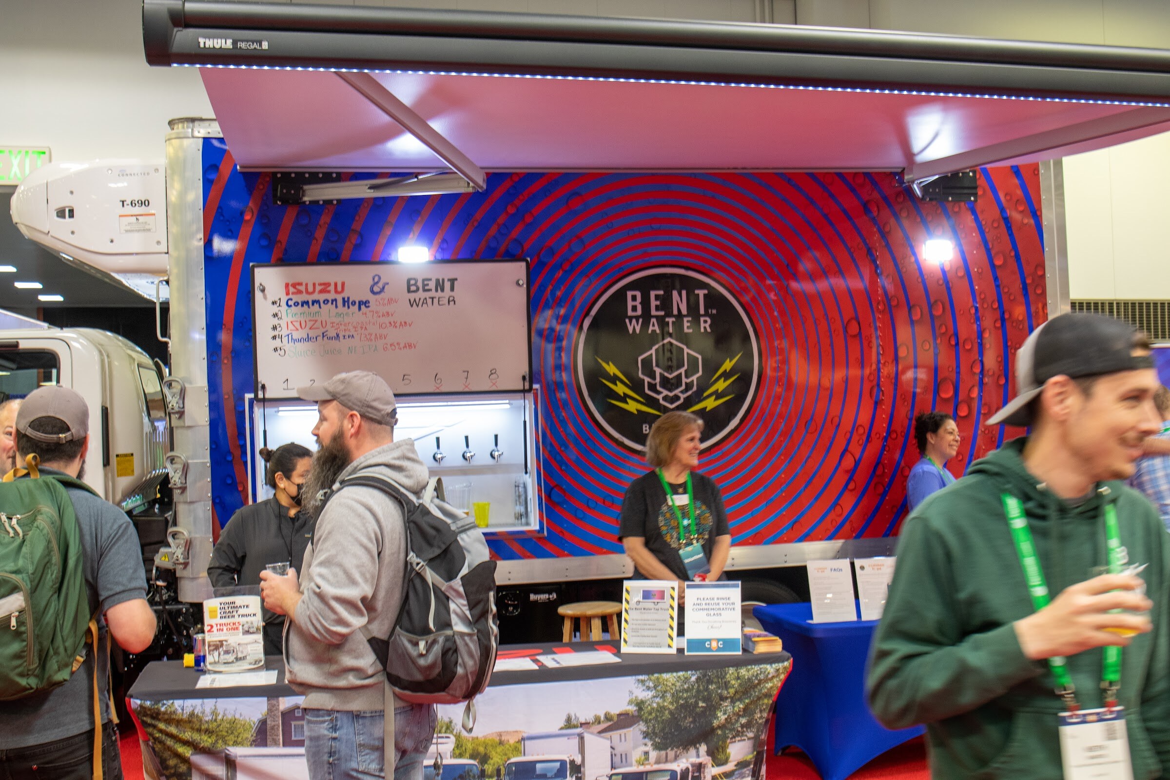 Booth staff stand behind the counter of a red and blue Bent Water display with a mounted tap tower