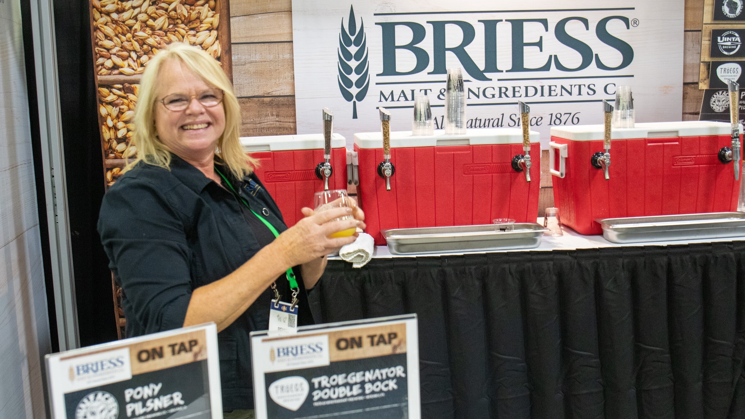A smiling woman holds a glass of beer in front of a Briess Malt and Ingredients booth with tap handles and On Tap signs