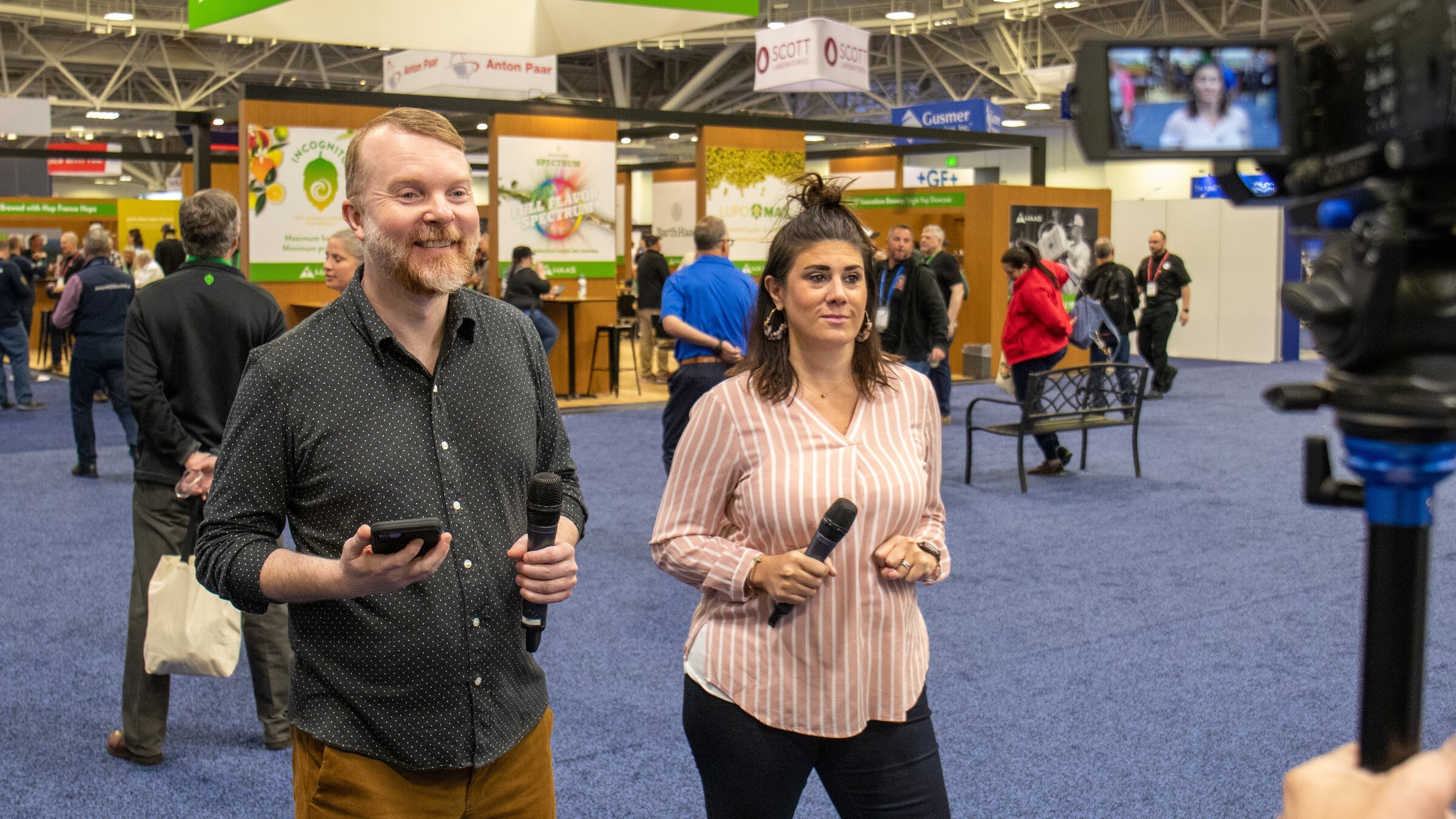 A man and a woman hold microphones and walk toward the camera on the show floor while attendees pass behind them
