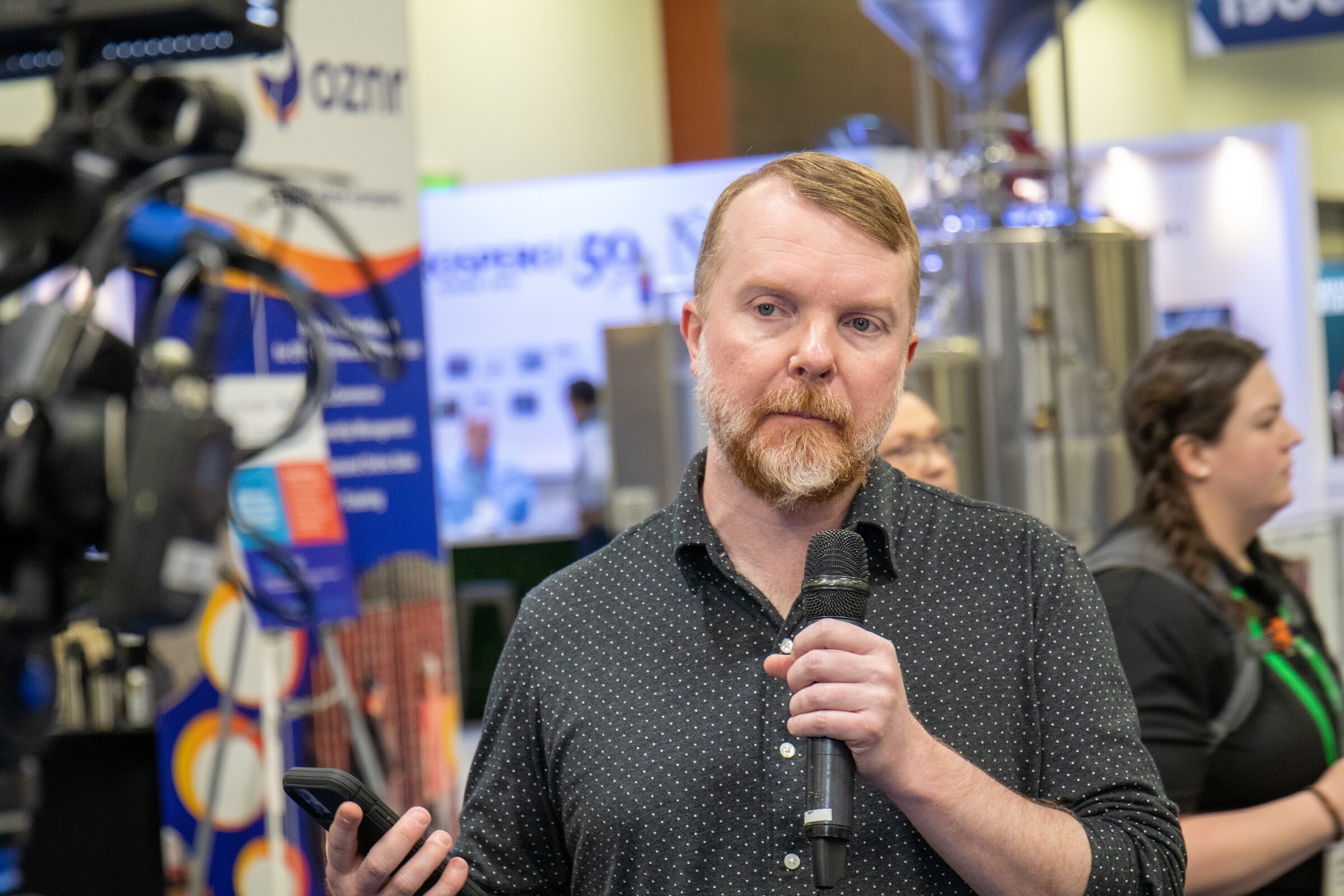 A bearded man in a dark patterned shirt holds a microphone and speaks to the camera in front of brewing equipment booths