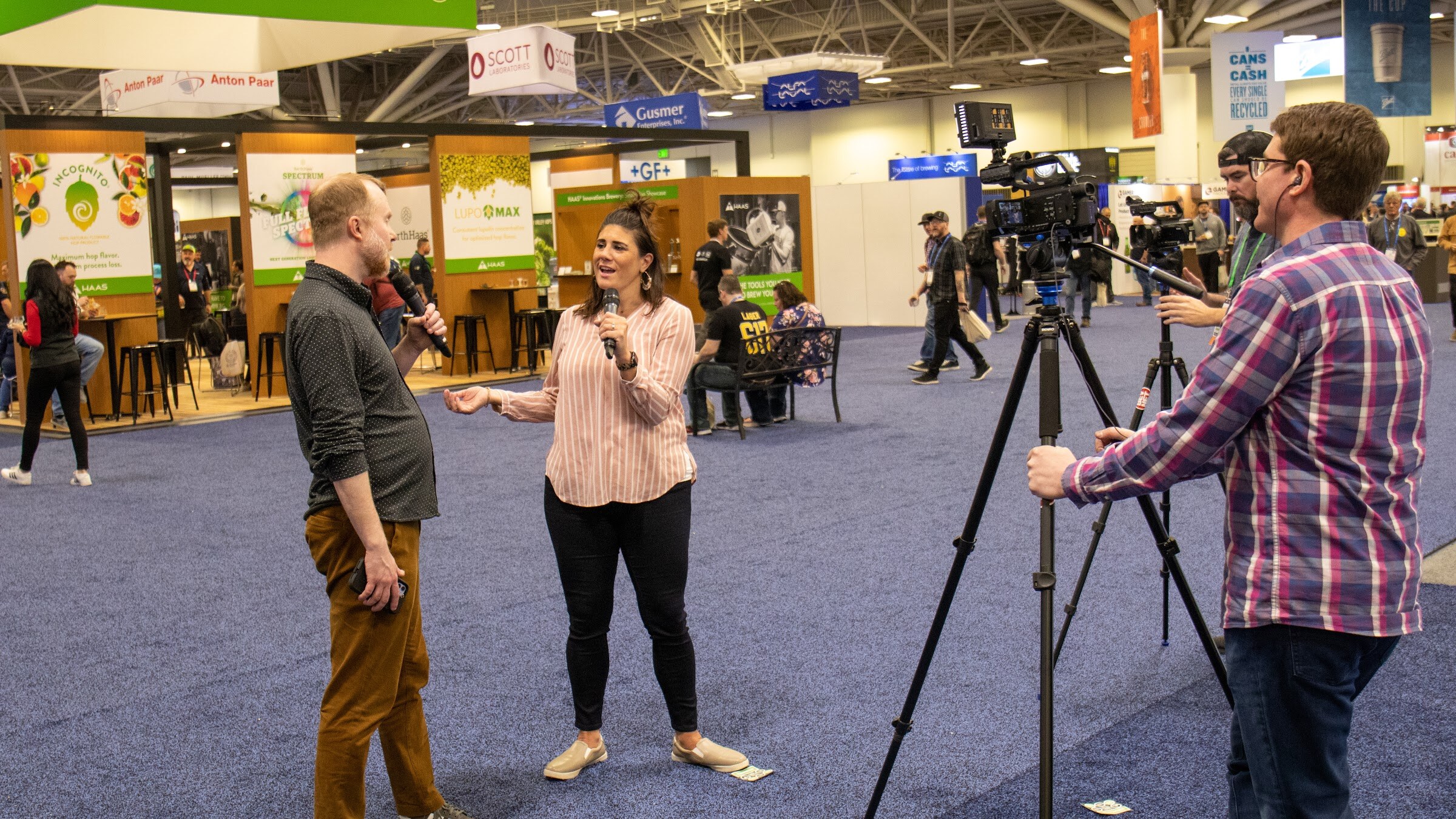 Two hosts with microphones stand in front of a camera operator and tripod in an aisle of the BrewExpo America show floor