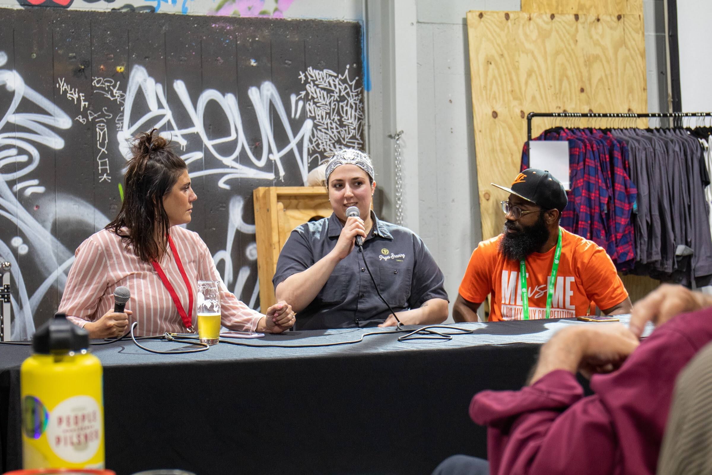 Three panelists with microphones sit at a draped table in front of a graffiti wall with a pint of beer, as audience members listen in the foreground