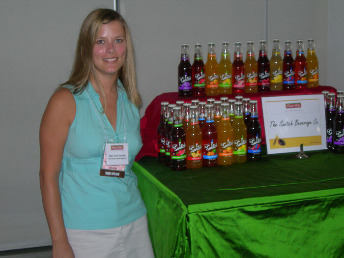 Mary Beth Bowen of The Switch Beverage Co., in a teal sleeveless top, stands beside a green-draped table displaying rows of Switch bottles and a sign reading The Switch Beverage Co.
