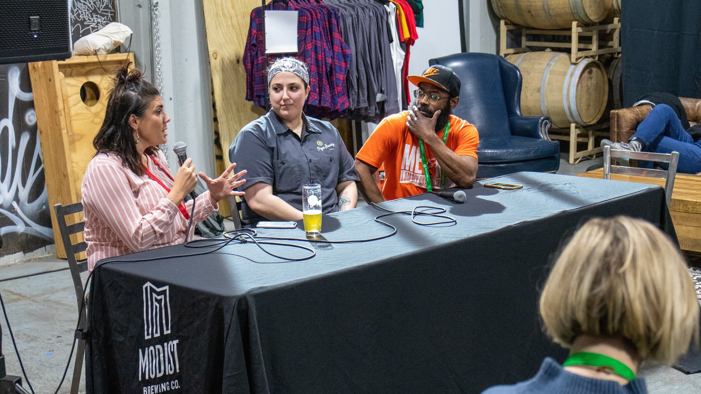 A woman speaks and gestures to two panelists seated at a black table cloth printed with the Modist Brewing Co. logo in a brewery with barrels behind