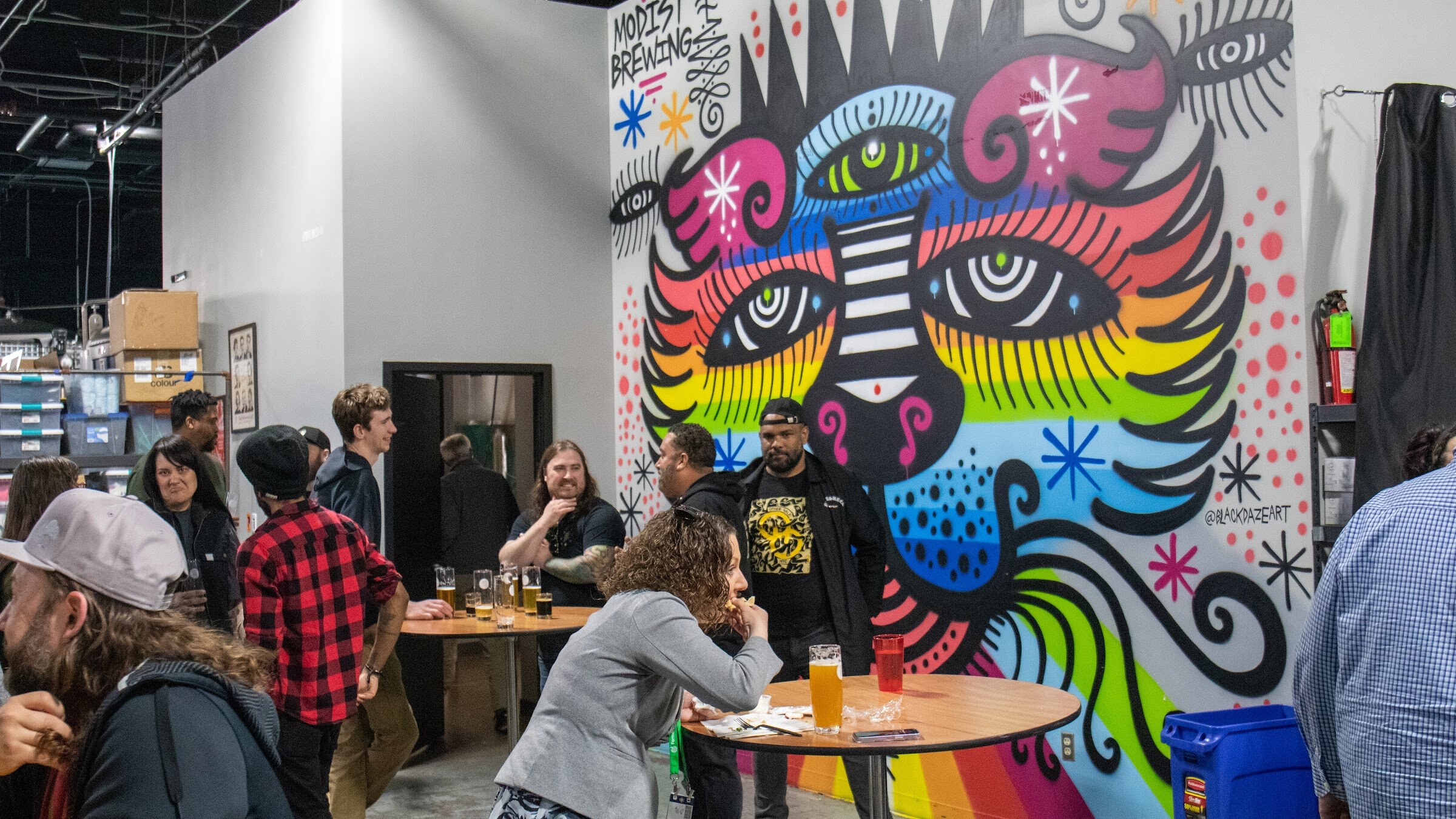 Guests talk around a tall table in front of a colorful mural of a face with rainbow stripes in a brewery taproom