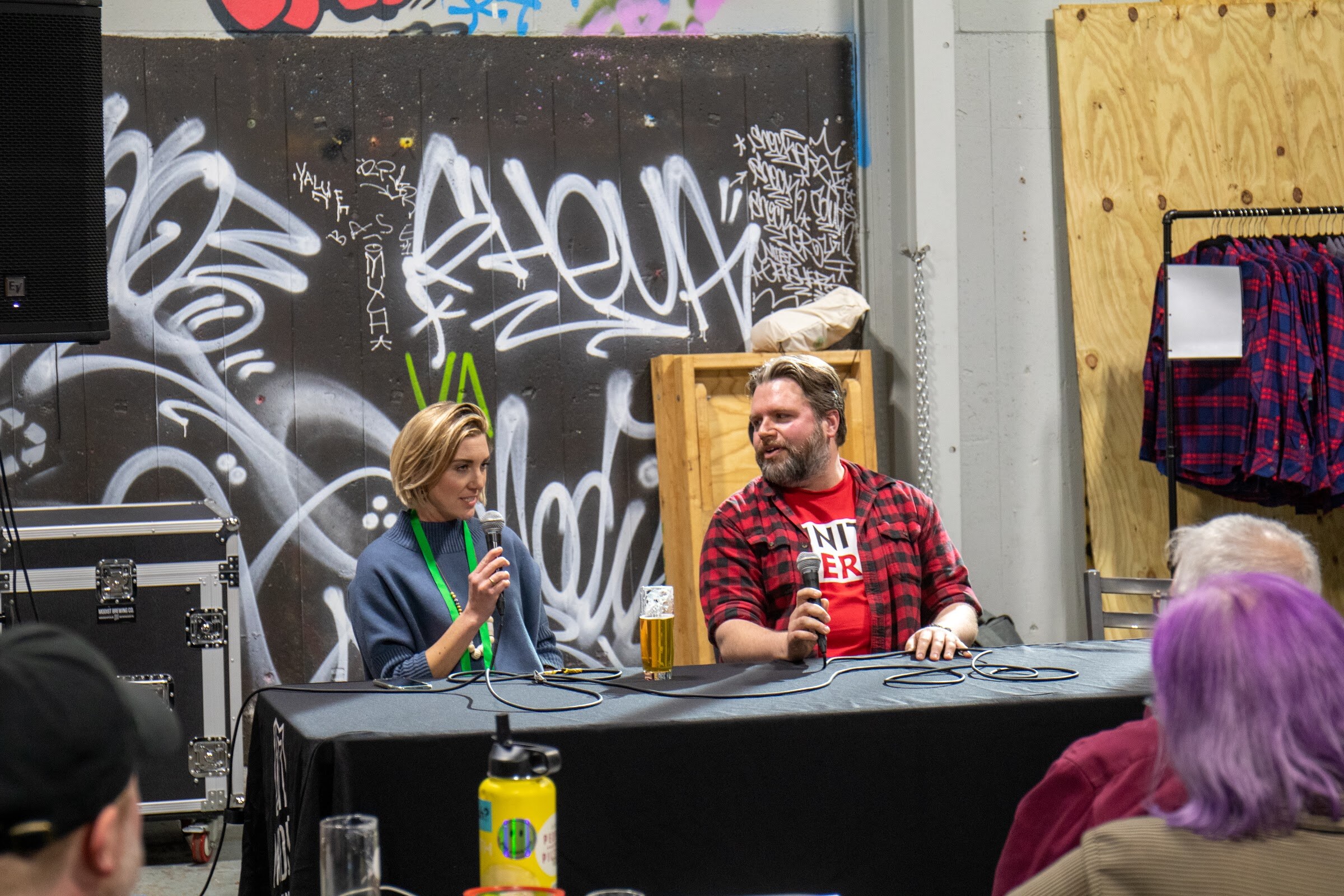 A woman in a blue sweater and a bearded man in a red plaid shirt hold microphones and talk at a draped table in front of a graffiti wall