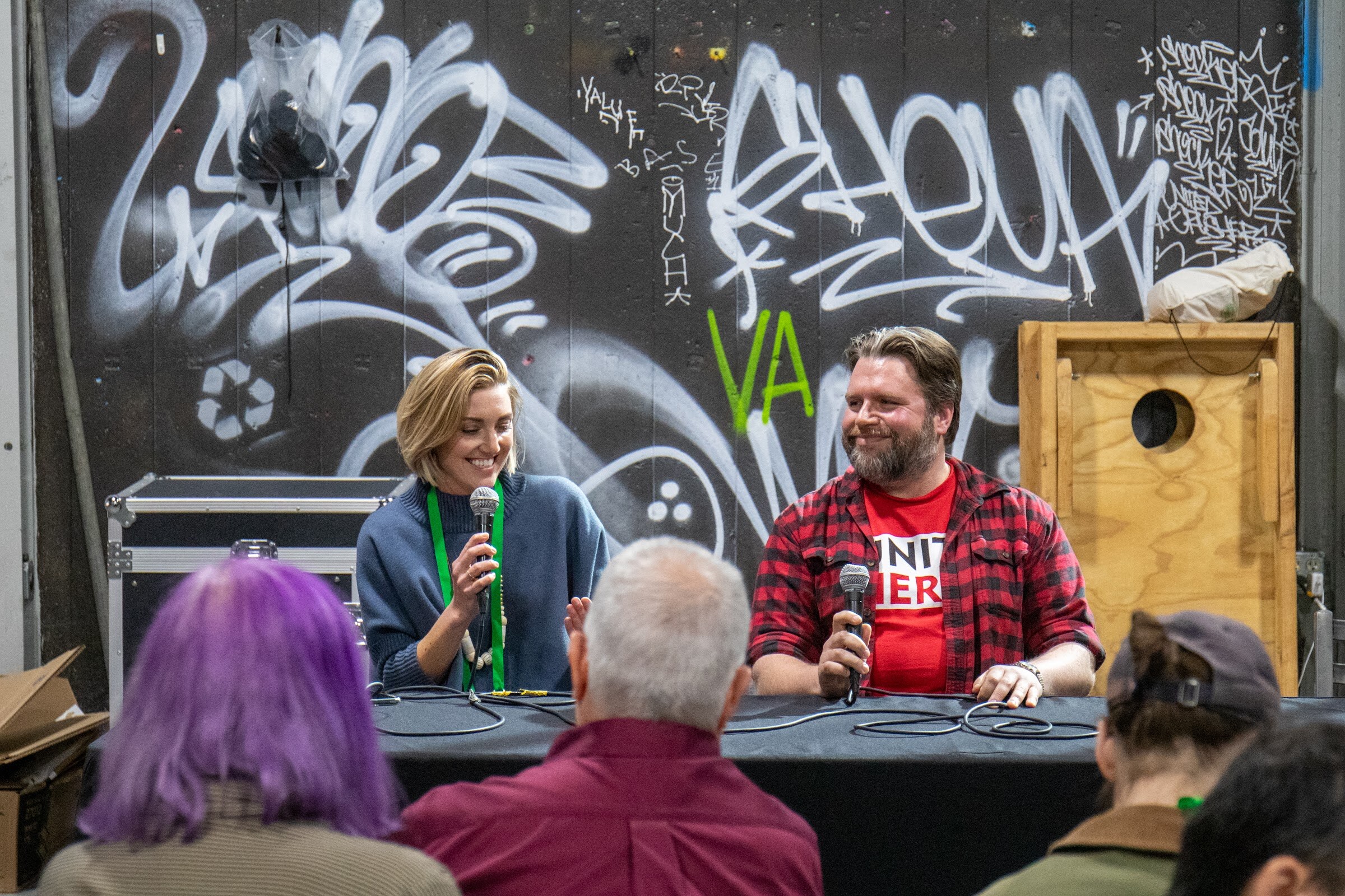Seated audience members face two speakers with microphones at a draped table in front of a graffiti wall and a wooden box