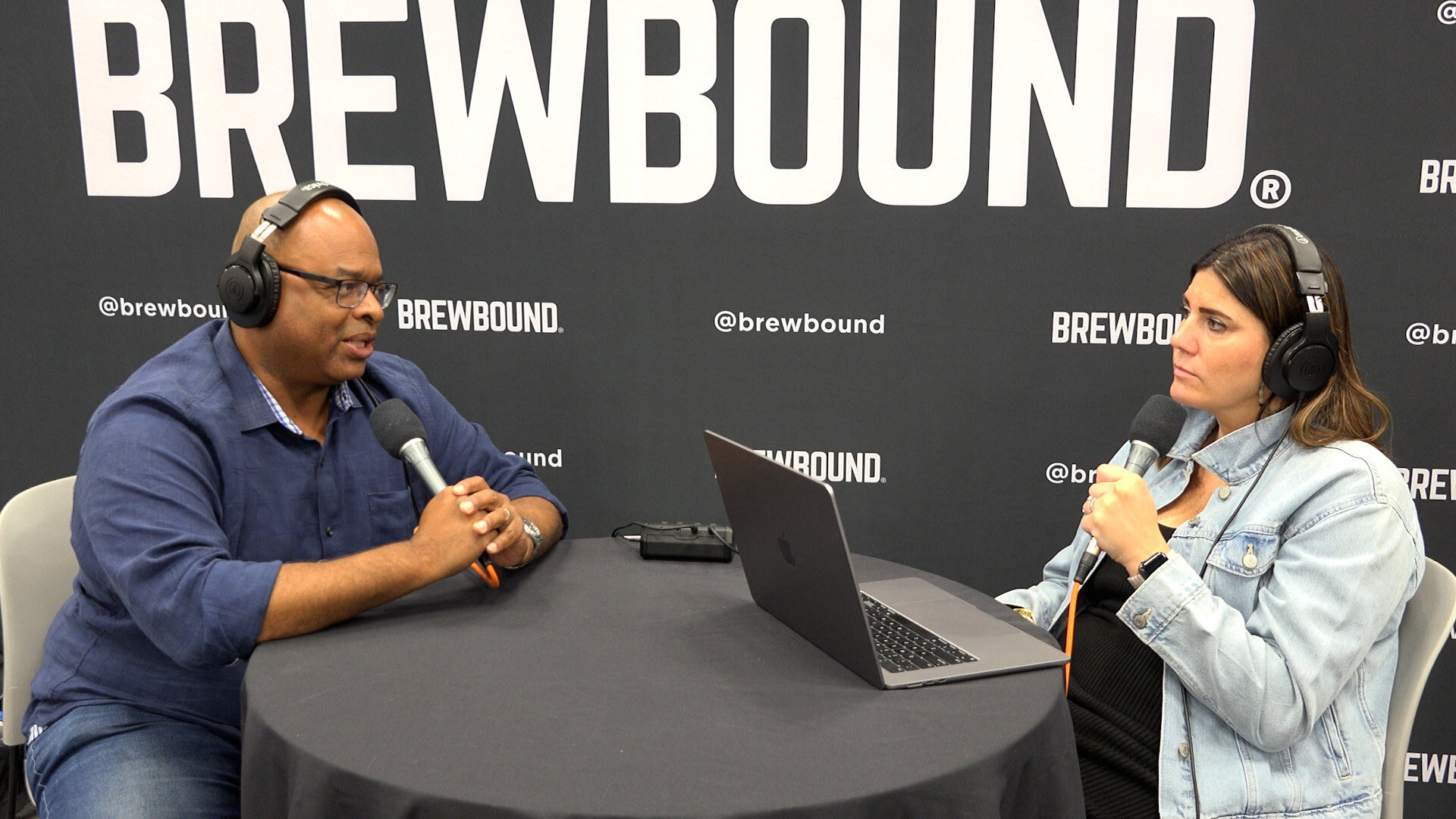 Man in a blue shirt and headphones talking with a woman in a denim jacket at a round table in front of a Brewbound backdrop.