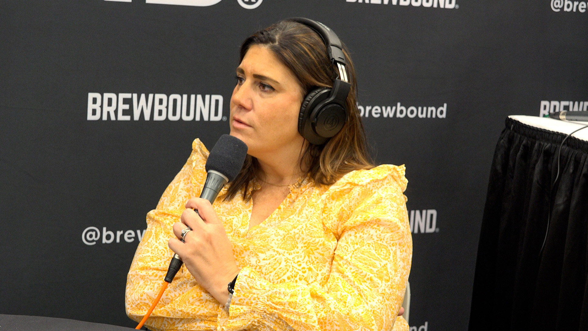 Woman in a yellow floral top and headphones holding a microphone in front of a Brewbound backdrop.
