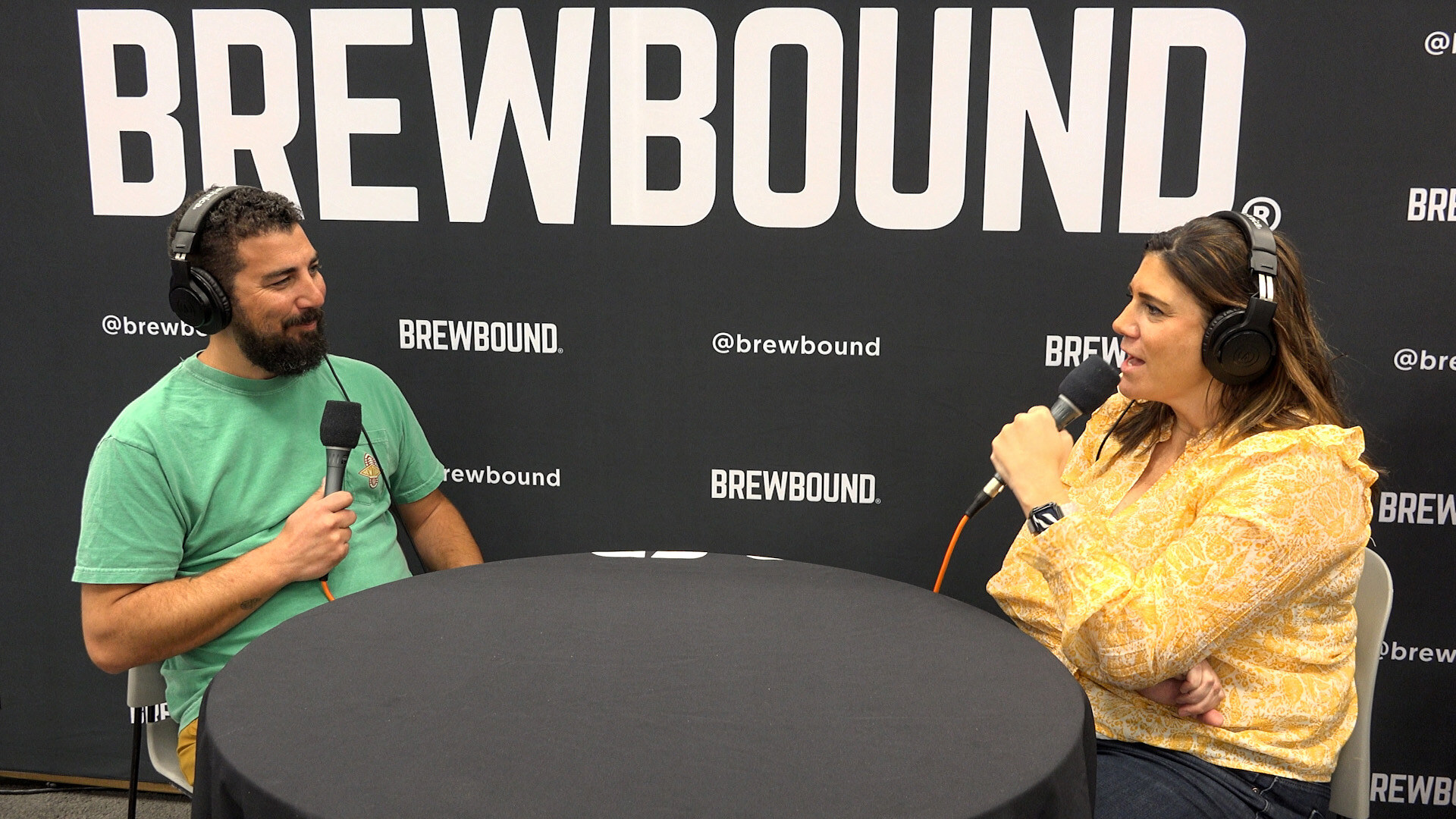 Man in a green T-shirt and headphones seated across a round table from a woman in a yellow top.