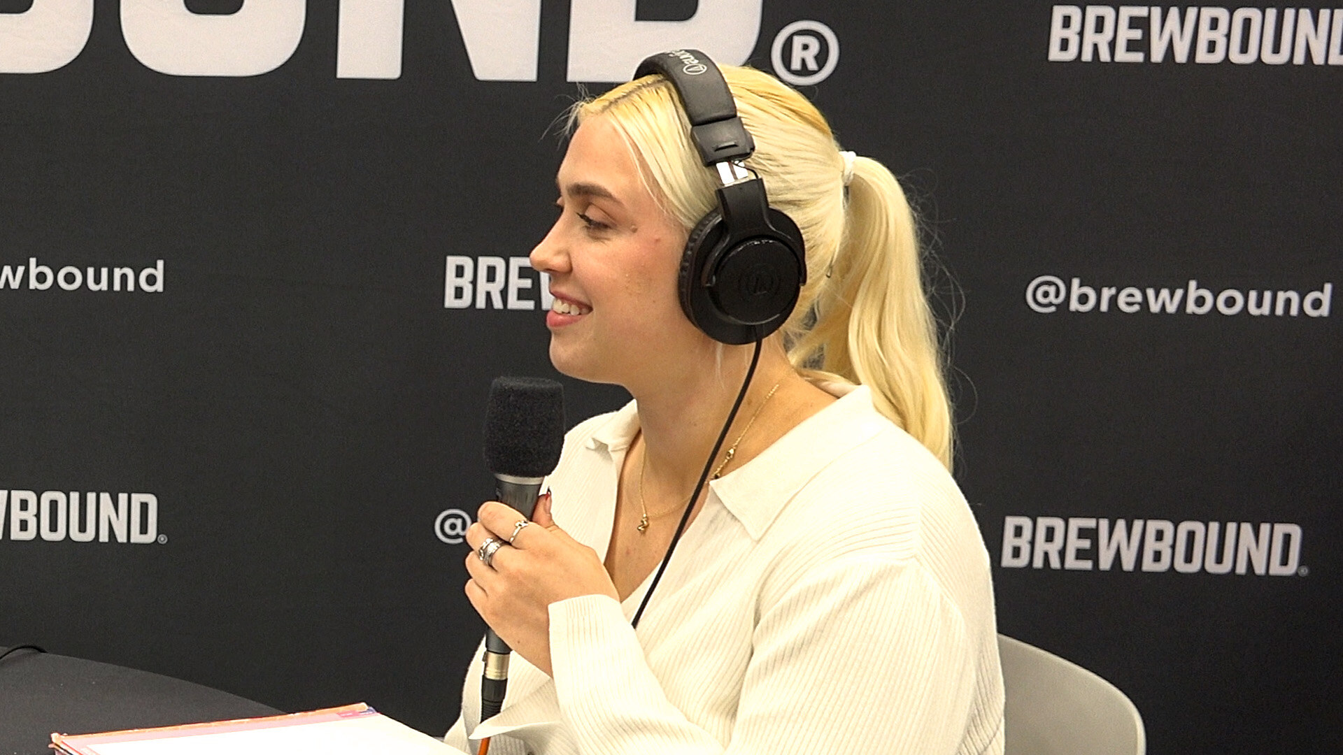 Woman with blonde ponytail and headphones holding a microphone in front of a Brewbound backdrop.