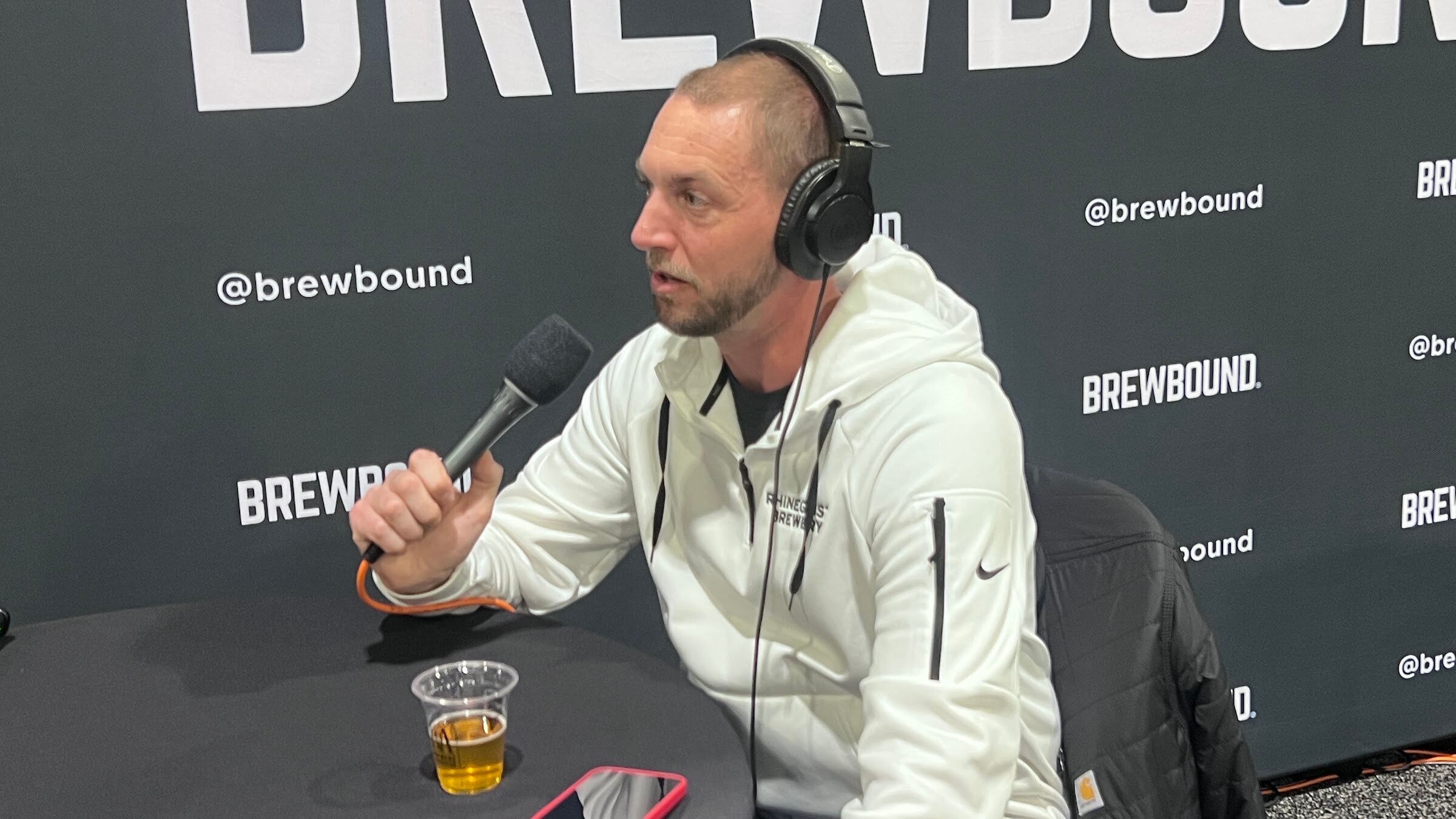 Man in a white hoodie and headphones speaks into a microphone at a table with a beer, in front of the Brewbound backdrop.