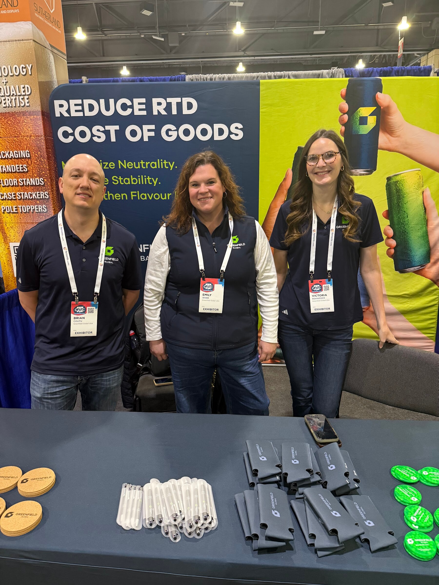 Three people in matching navy shirts pose in front of a Greenfield Global banner reading Reduce RTD cost of goods.