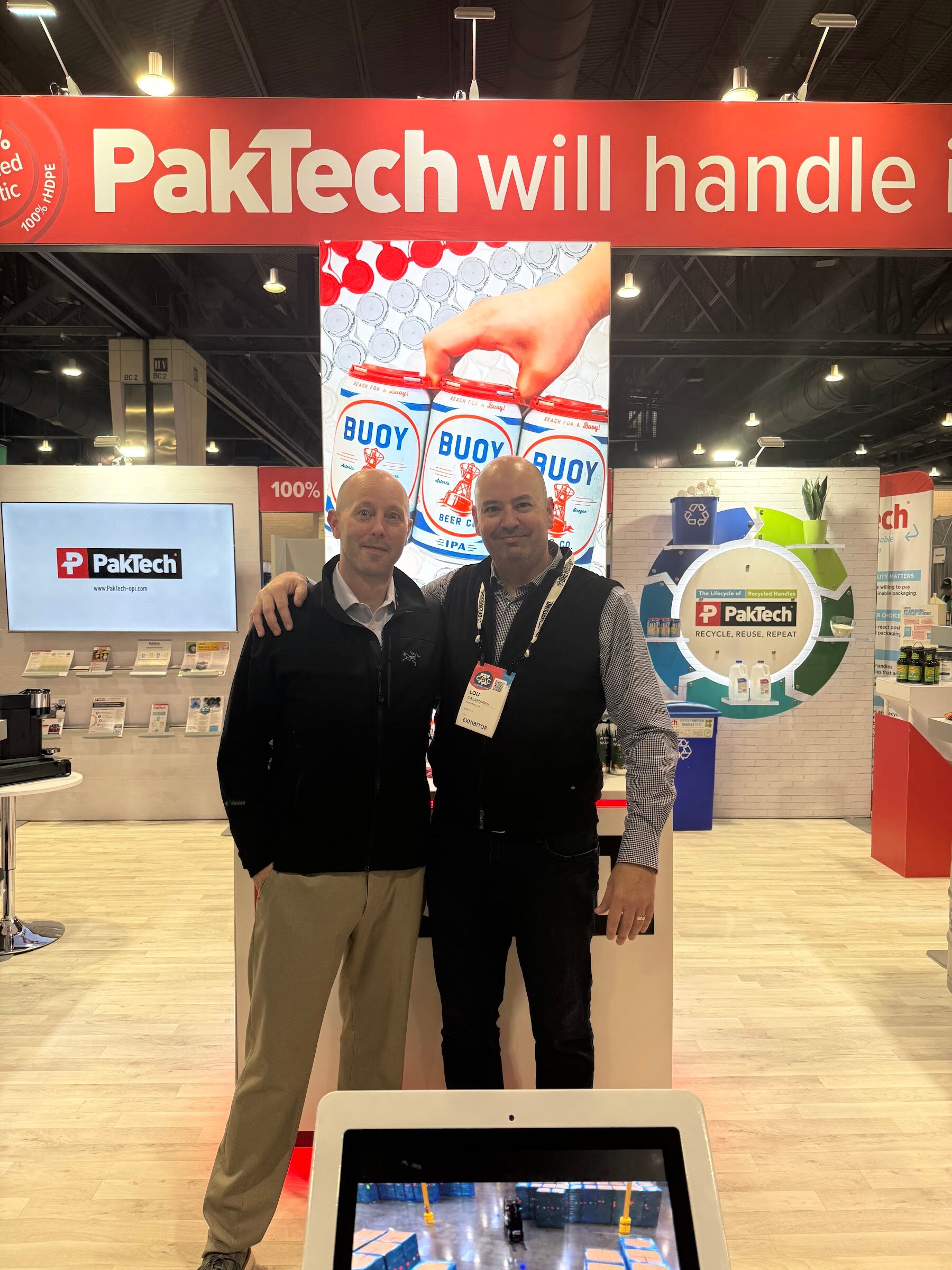 Two men pose under a red PakTech will handle it sign beside a display of can carriers.