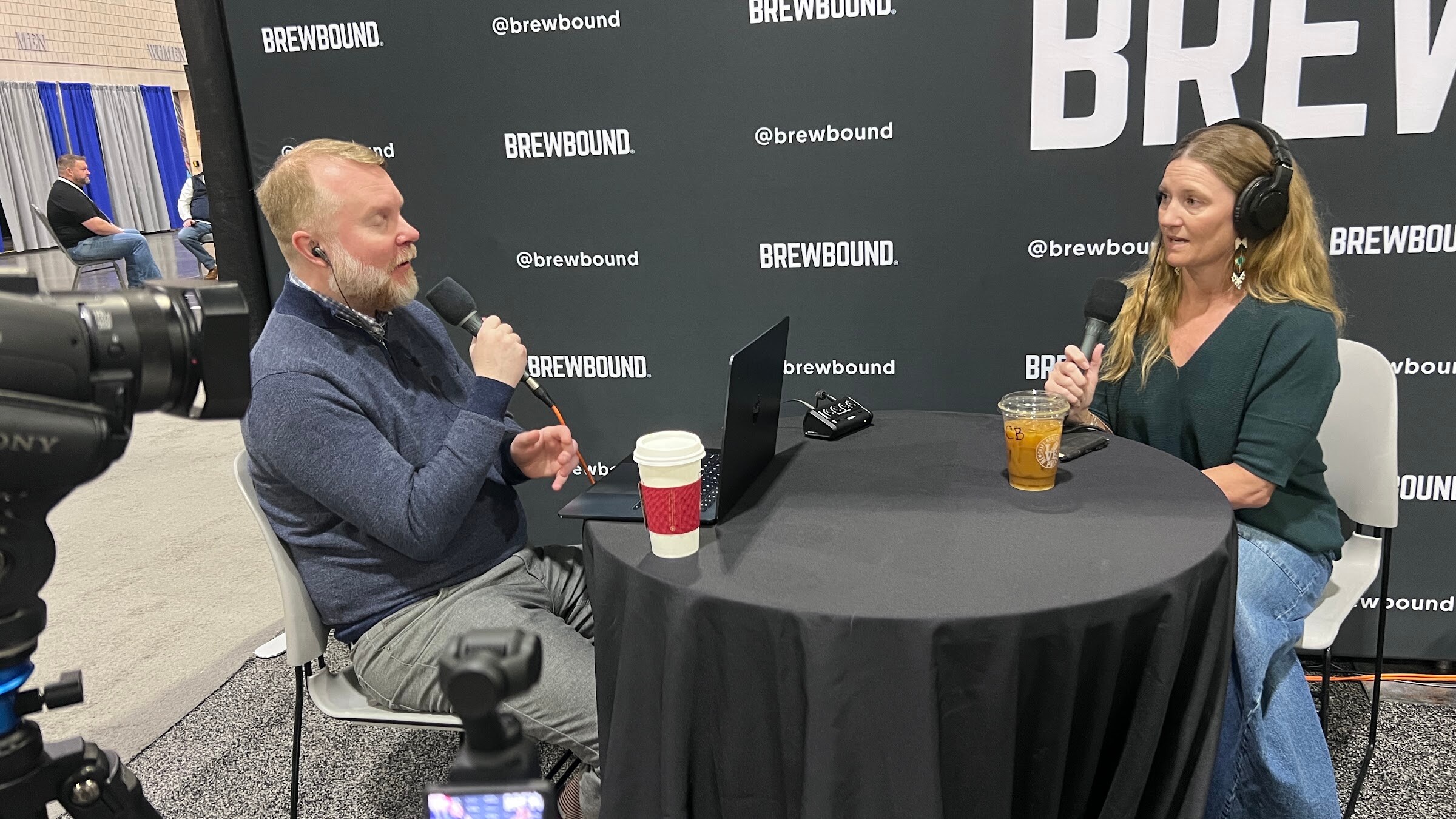 Man and woman in headphones talk into microphones across a round table with a laptop and drinks, a video camera in the foreground.