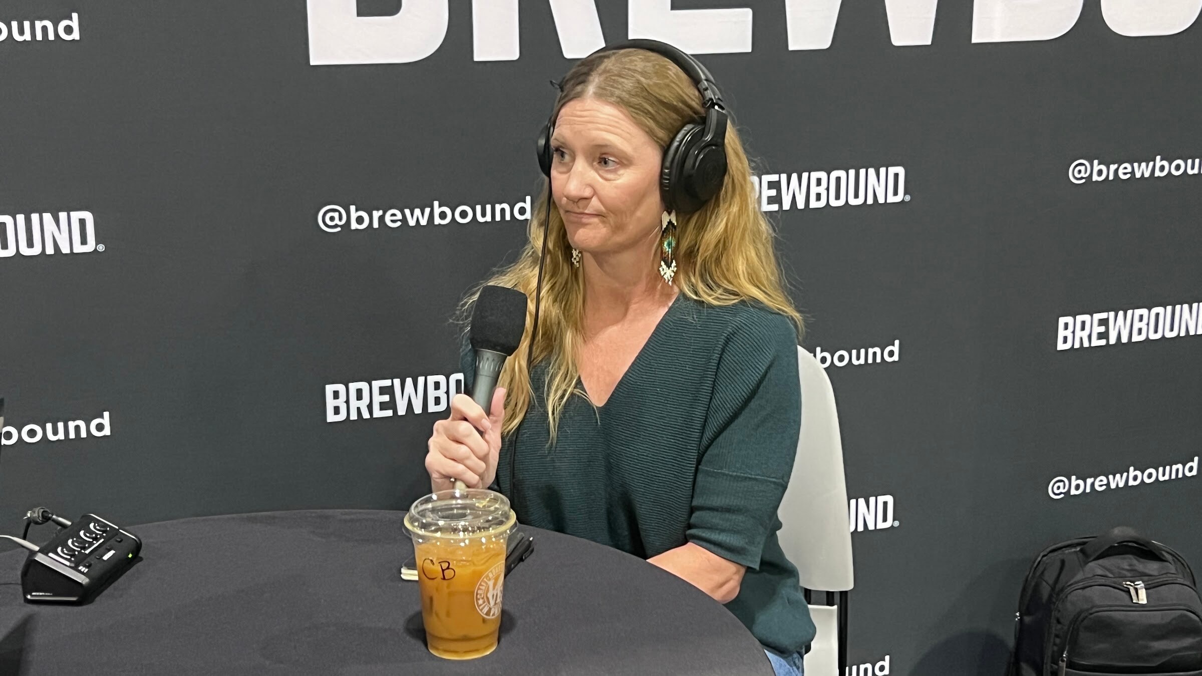 Woman with long hair in headphones holds a microphone beside an iced coffee in front of the Brewbound backdrop.