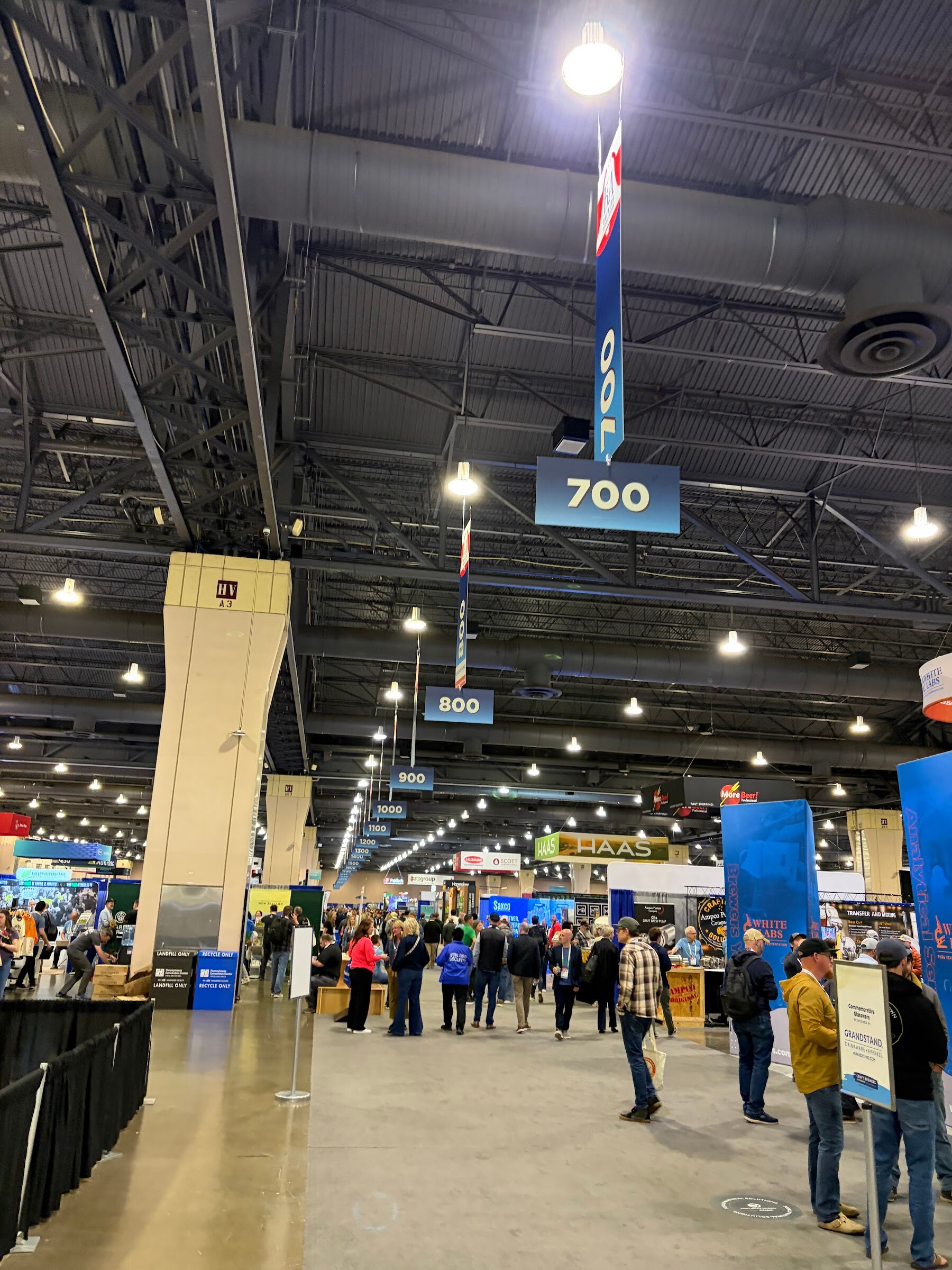 Wide aisle of the BrewExpo America trade show floor with attendees, booth signs and hanging aisle markers.