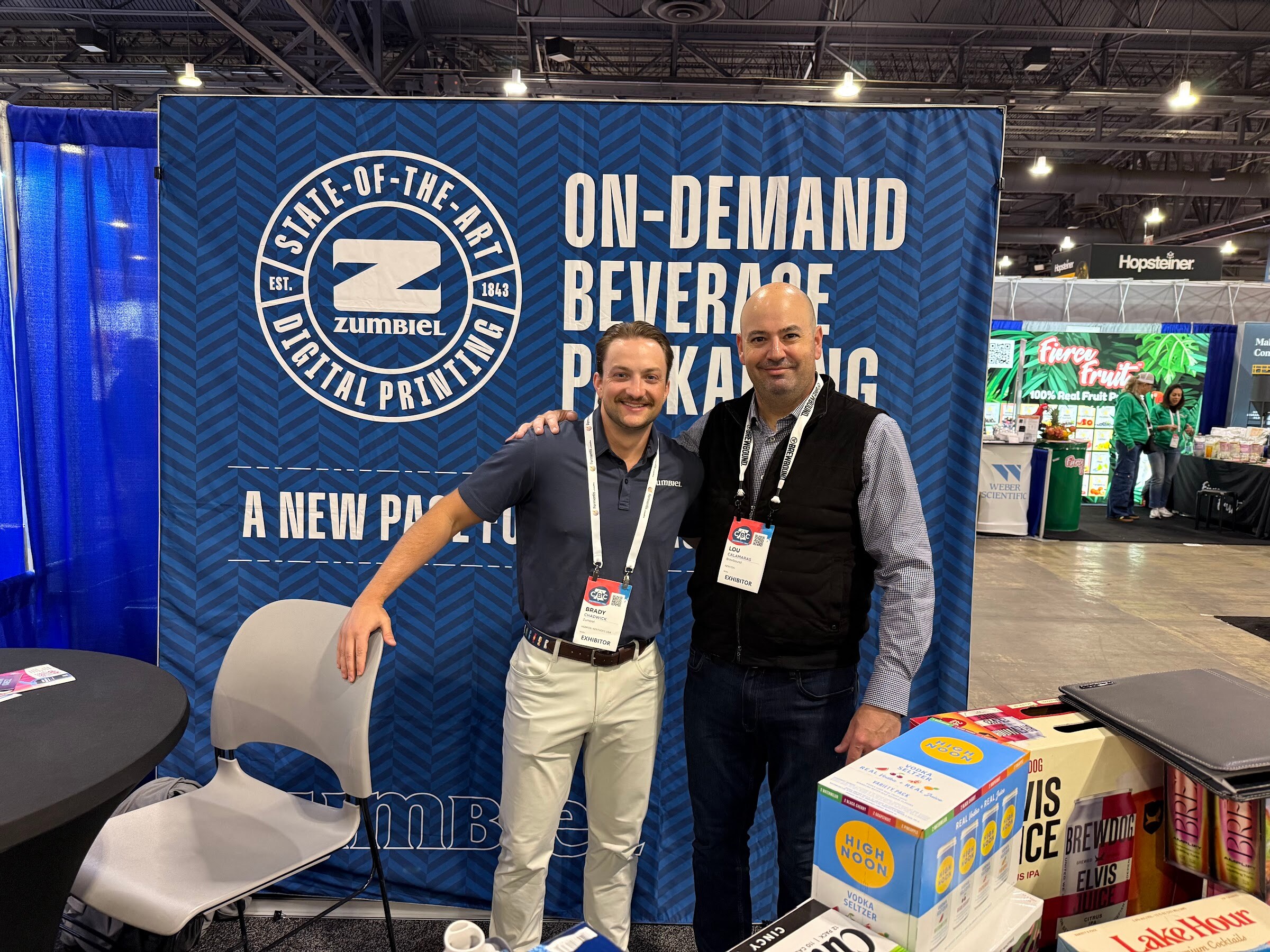 Two men pose in front of a blue Zumbiel banner reading On-Demand Beverage Packaging, beside printed beer and seltzer cartons.