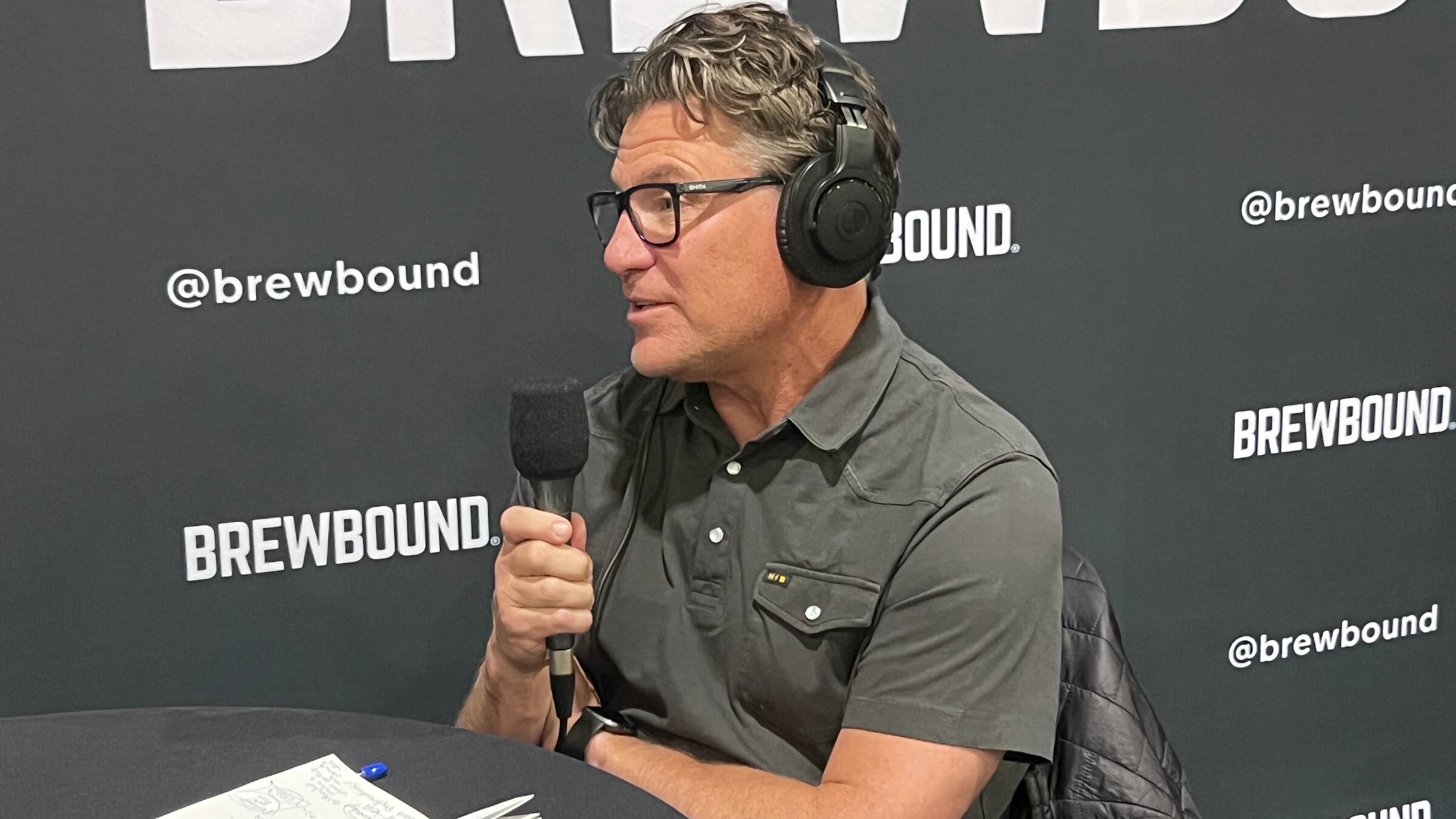 Man in glasses, headphones and a gray shirt speaks into a microphone in front of the Brewbound backdrop.