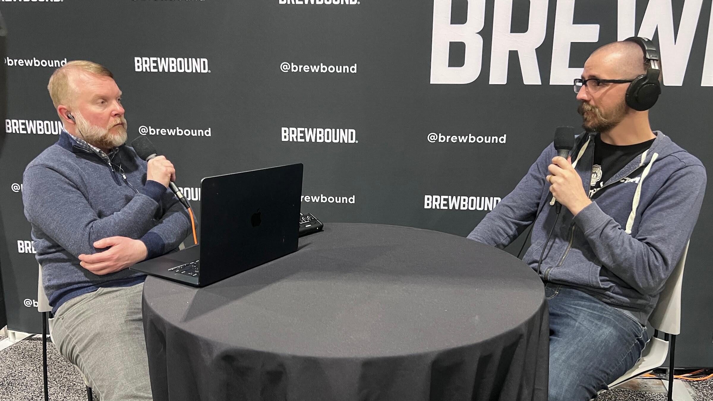 Two men in headphones with microphones sit at a round table with a laptop under a large Brewbound wordmark.