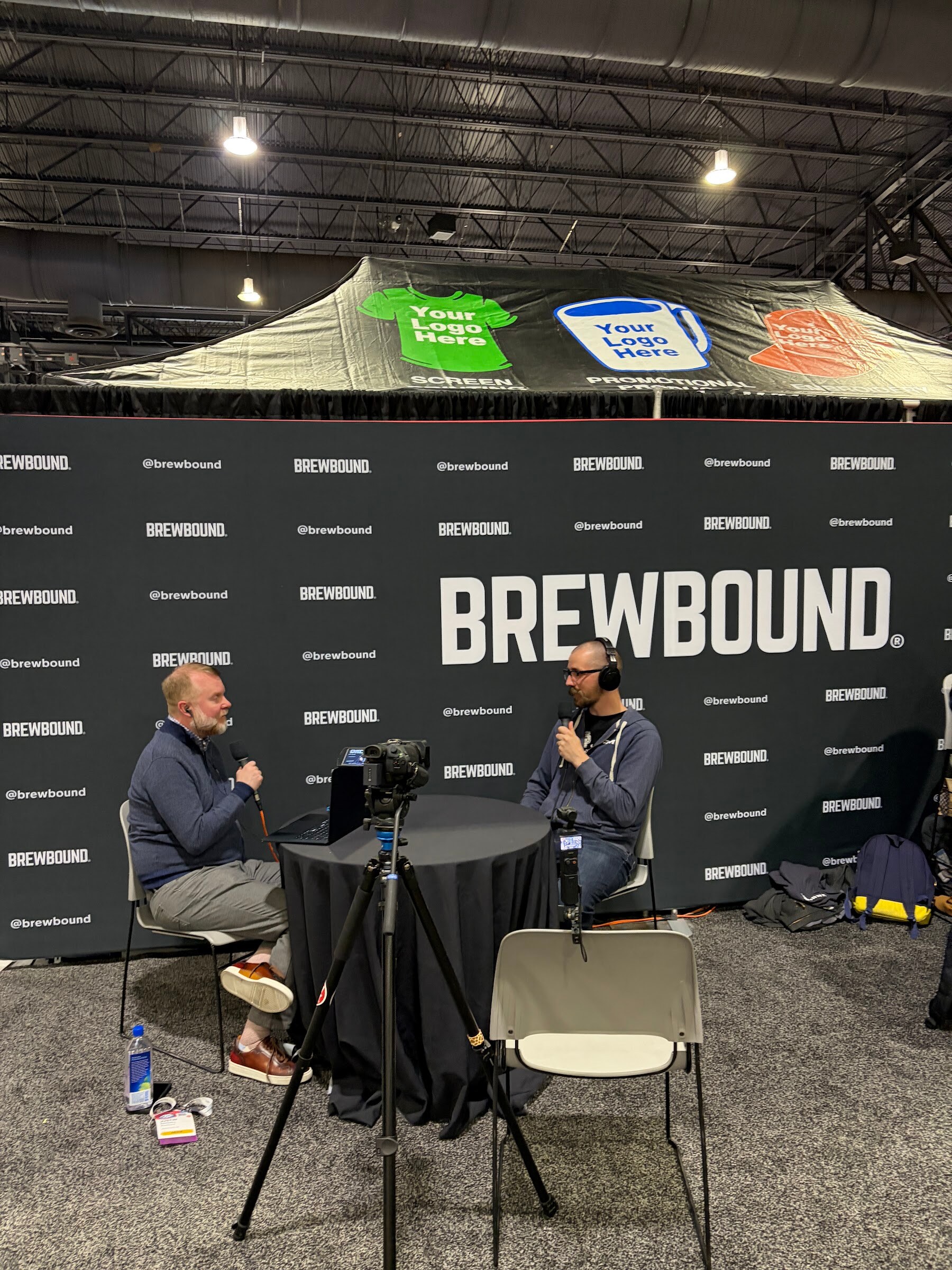 Two people talk into microphones at a round table in front of a large Brewbound backdrop, with a camera on a tripod in the foreground.
