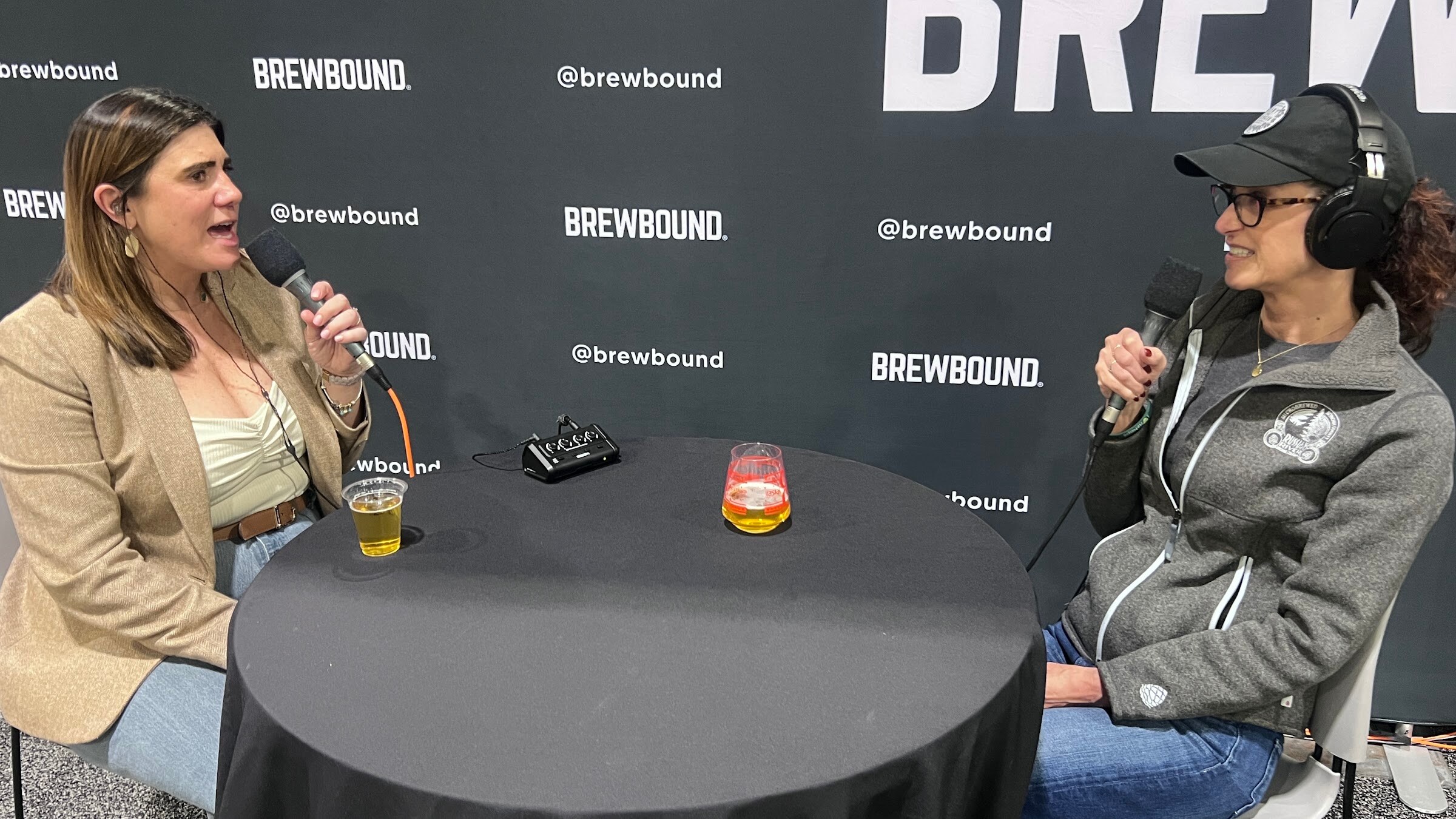 Two women in headphones talk into microphones across a round table with two glasses of beer in front of the Brewbound backdrop.
