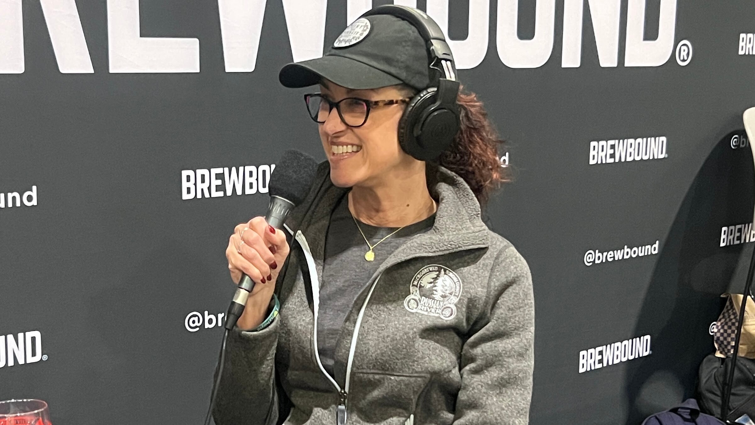 Smiling woman in a cap, glasses and headphones holds a microphone in front of the Brewbound backdrop.