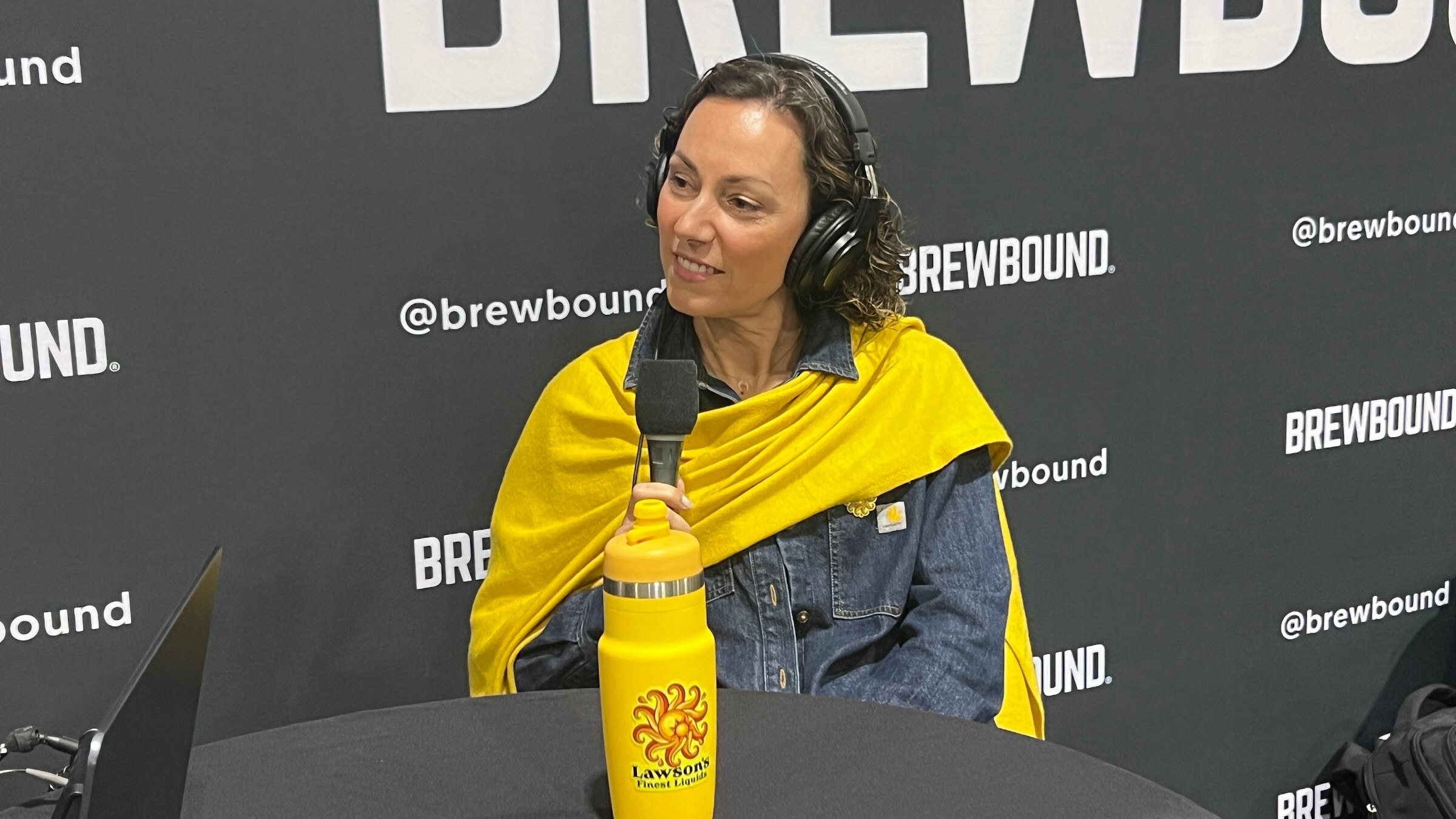 Woman in headphones and a yellow scarf holds a microphone beside a yellow Lawson’s Finest Liquids water bottle.
