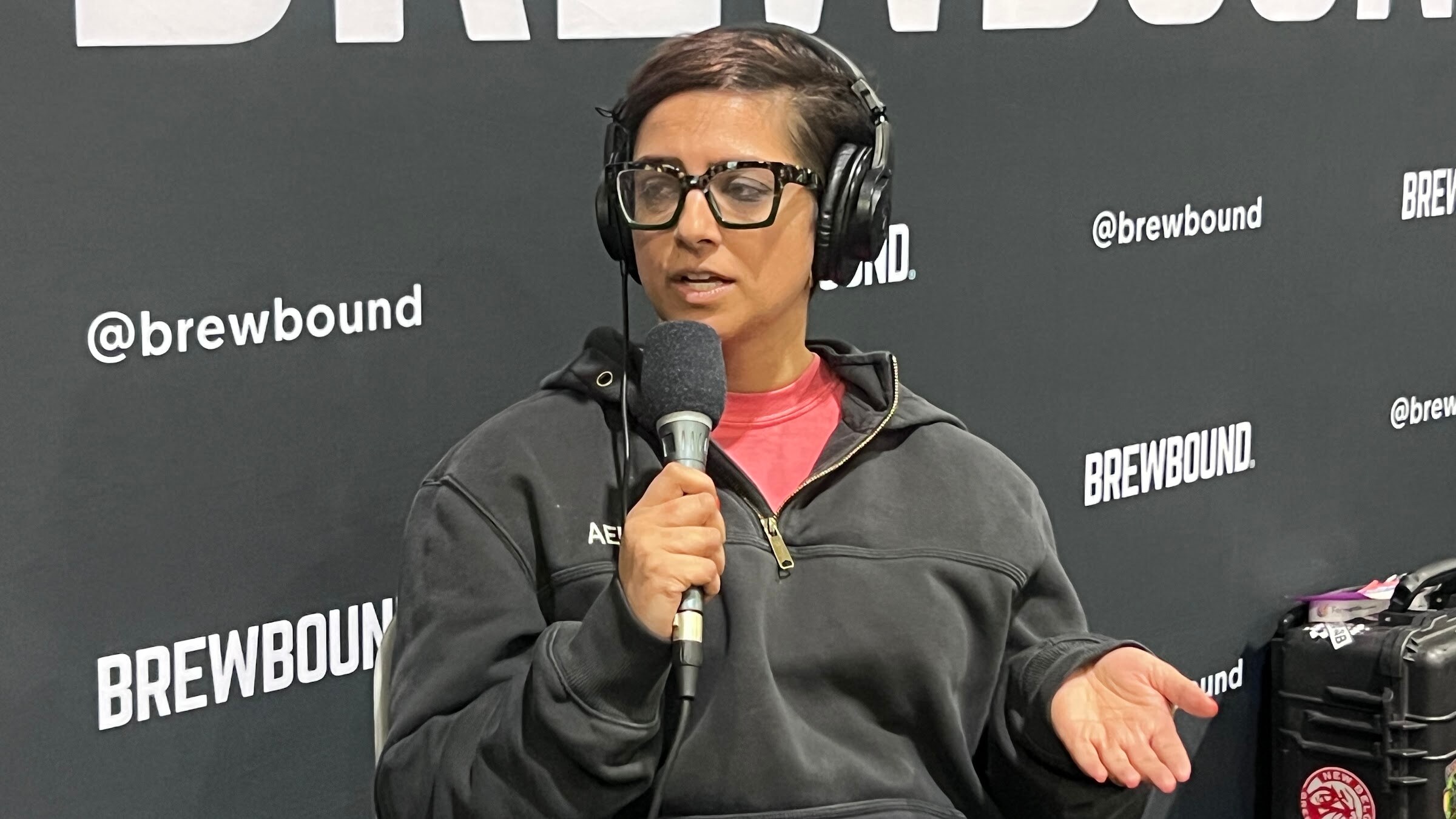 Woman in glasses, headphones and a dark hoodie gestures while speaking into a microphone in front of the Brewbound backdrop.
