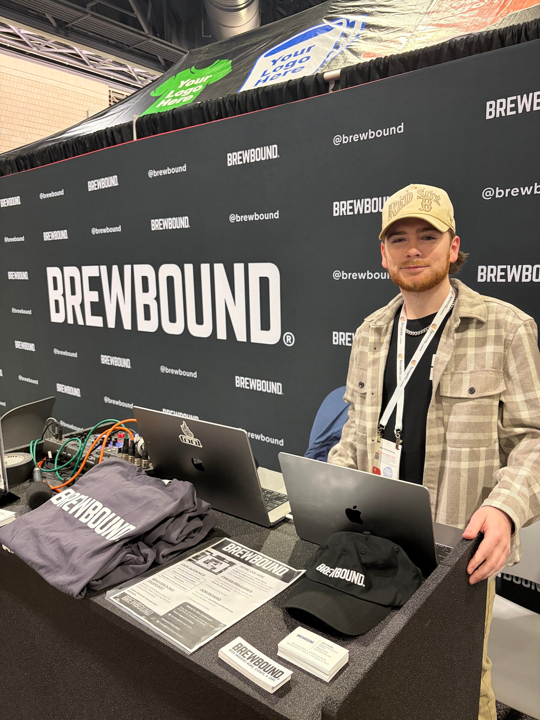 Smiling young man in a plaid jacket stands at a counter with laptops, Brewbound T-shirts and a Brewbound cap in front of the Brewbound backdrop.