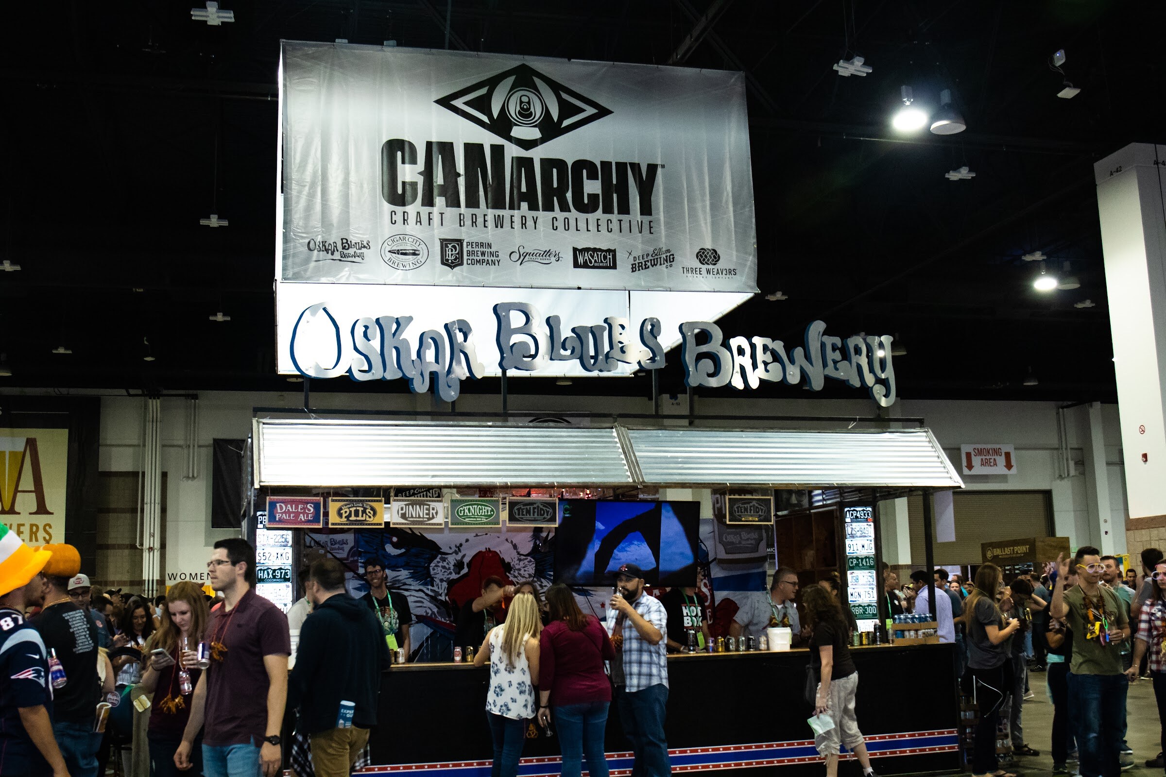 A large Canarchy Craft Brewery Collective banner above an Oskar Blues Brewery bar with taps, a crowd at the counter and a Patriots jersey in the foreground.