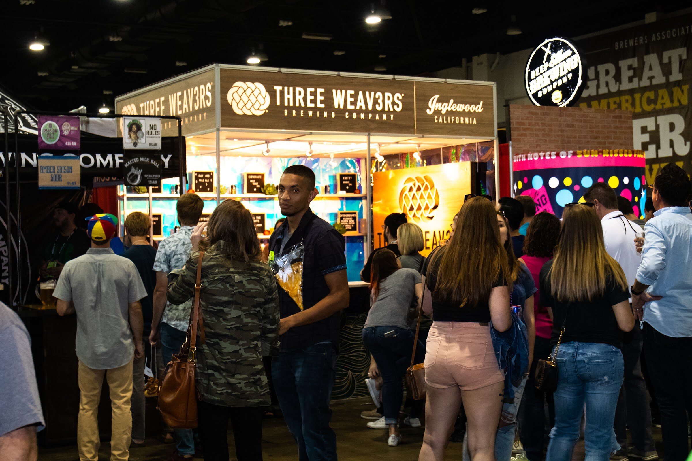 A lit Three Weavers Brewing Company booth with chalkboard menus and a line of attendees in front, with a Deep Ellum-style booth and the Great American Beer Festival sign at the upper right.