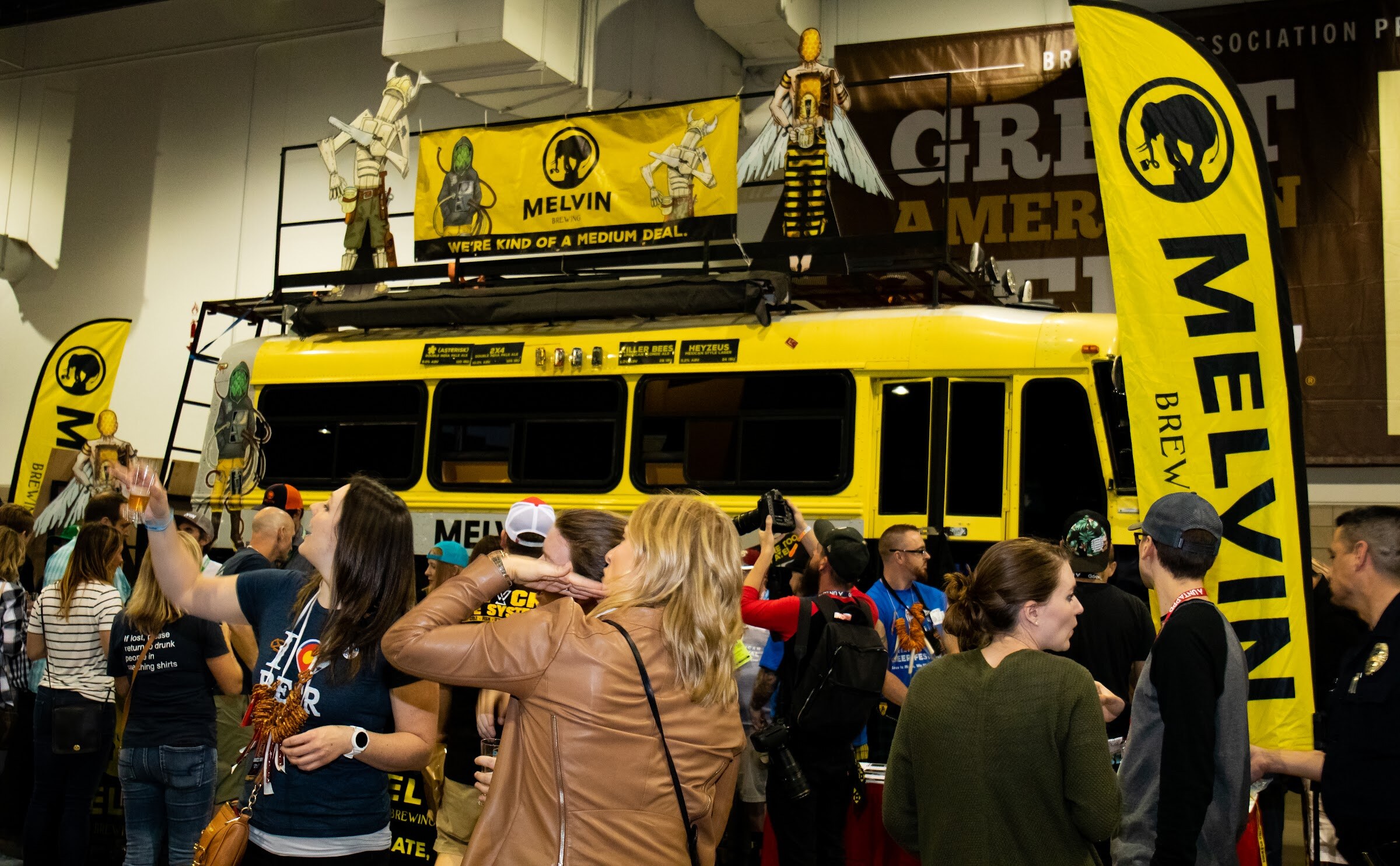 A yellow bus covered in Melvin Brewing banners and insect figures on its roof, with attendees in the foreground, one raising a glass.