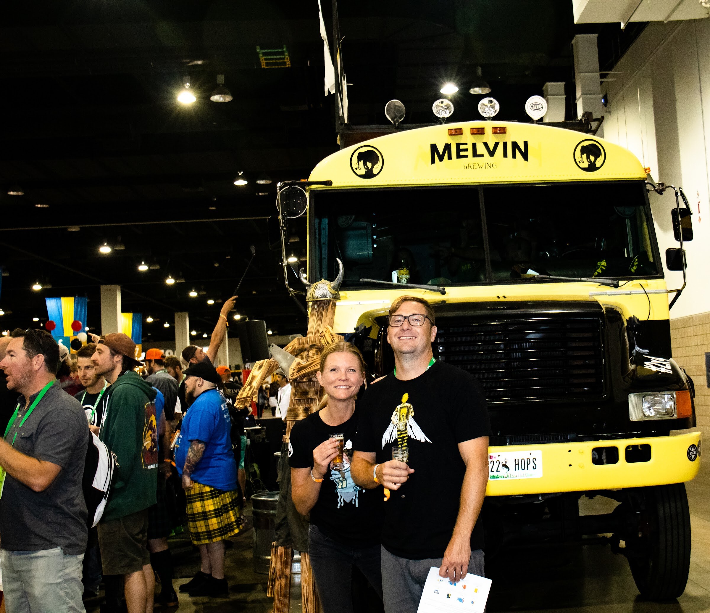 A man in glasses and a woman in black T-shirts hold tasters in front of the yellow Melvin Brewing bus with a 22 HOPS license plate, with festival-goers behind them.