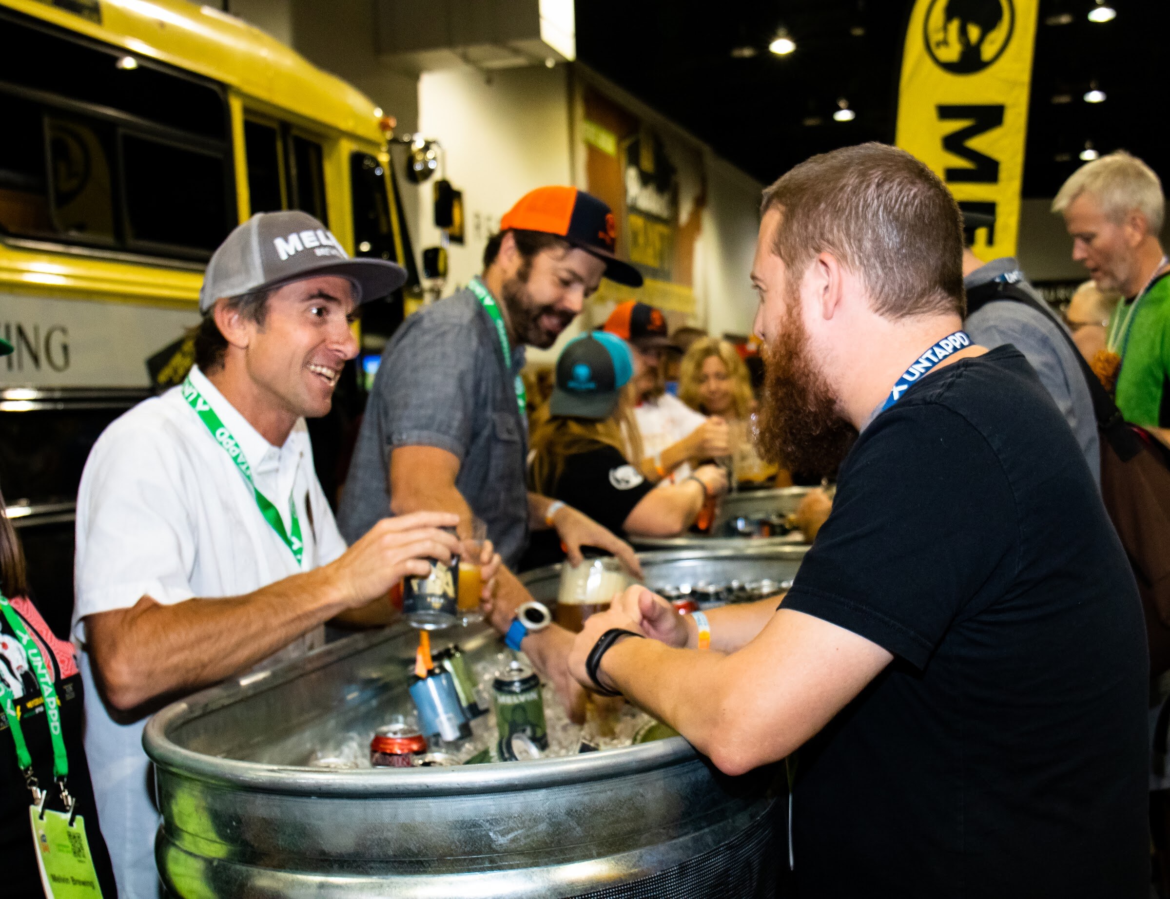 A smiling man in a Melvin cap and white shirt hands a beer across a metal tub of iced cans to a bearded festival-goer, with more staff in caps behind him.