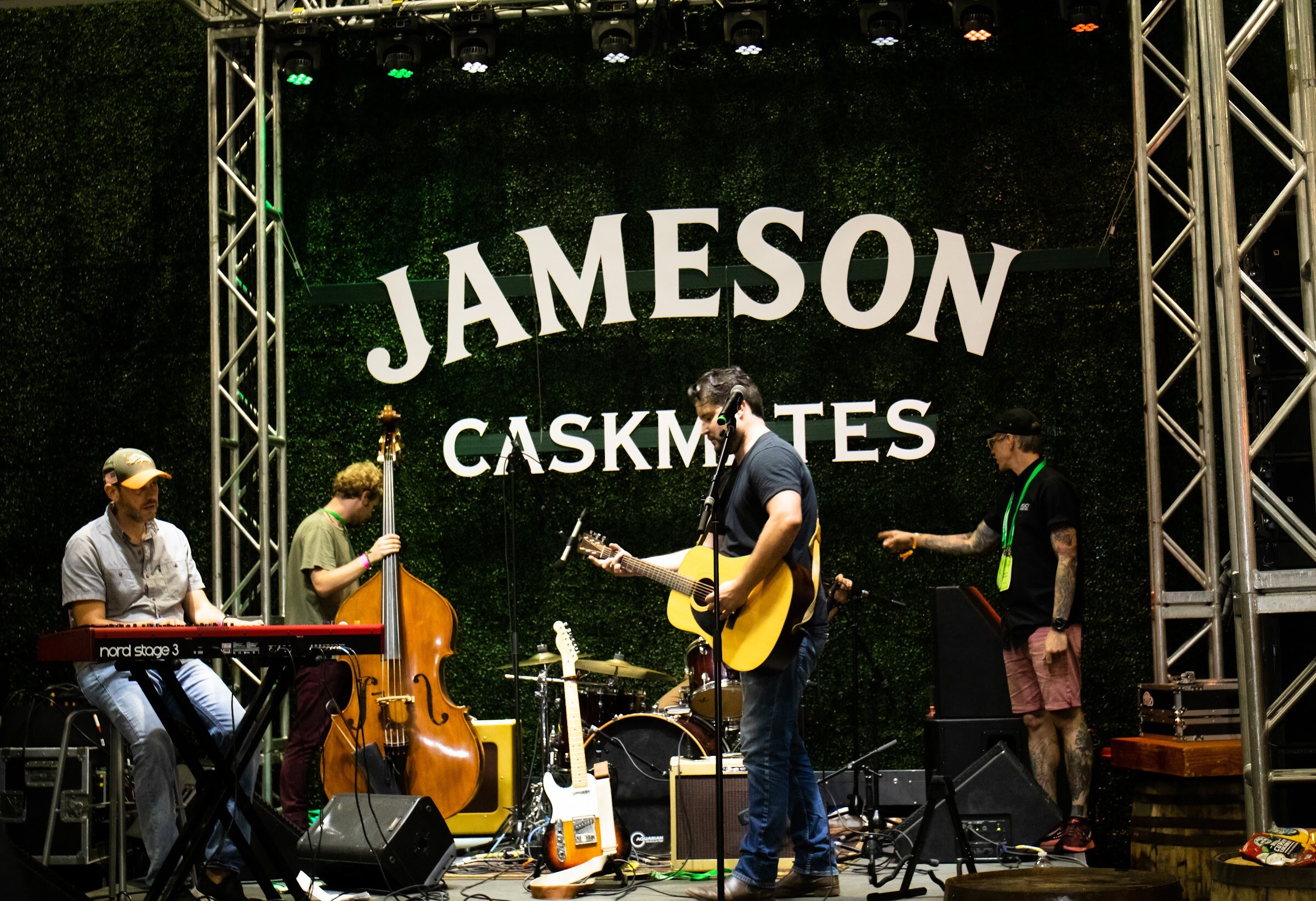 A keyboardist, an upright bassist, a guitarist and a technician on a stage with an ivy wall reading Jameson Caskmates.