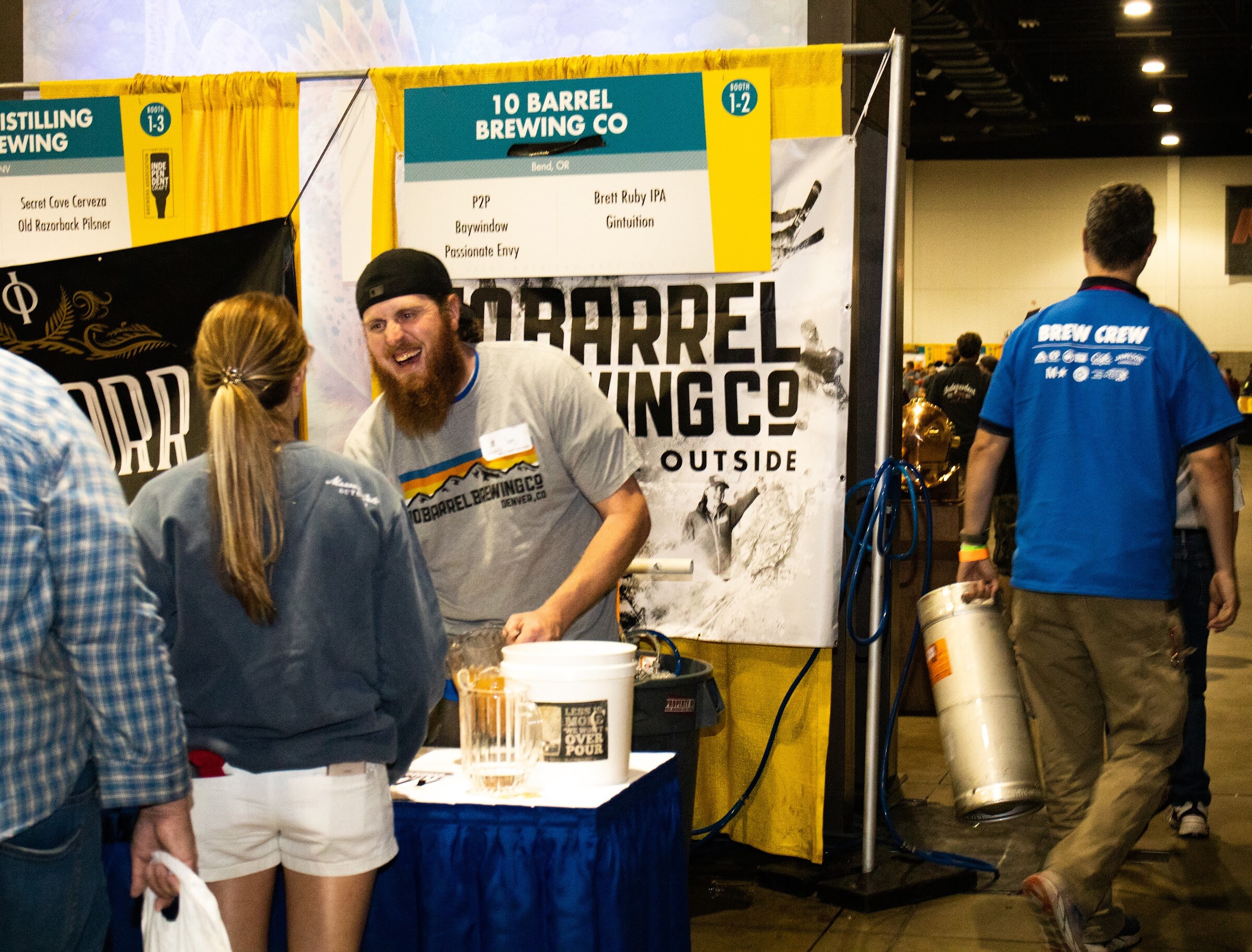 A bearded man in a 10 Barrel Brewing Co shirt leans over a blue-draped table at the 10 Barrel booth, with a woman in white shorts in front and a person in a blue Brew Crew shirt carrying a keg behind.