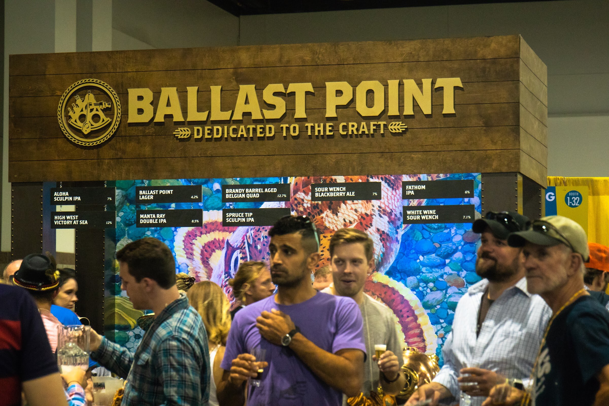A wooden Ballast Point Dedicated to the Craft sign over a mural and a list of beers including Sculpin IPAs, with attendees holding tasting glasses in the foreground.