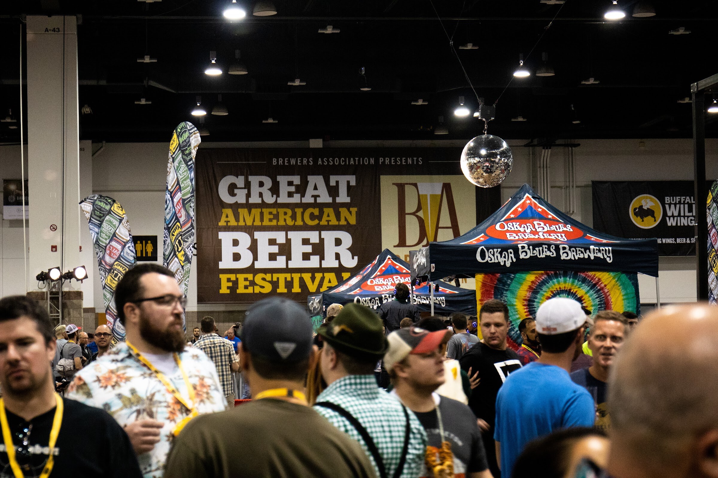 A brown Brewers Association Presents Great American Beer Festival banner above a crowd of attendees with lanyards, a disco ball and an Oskar Blues Brewery tent behind.