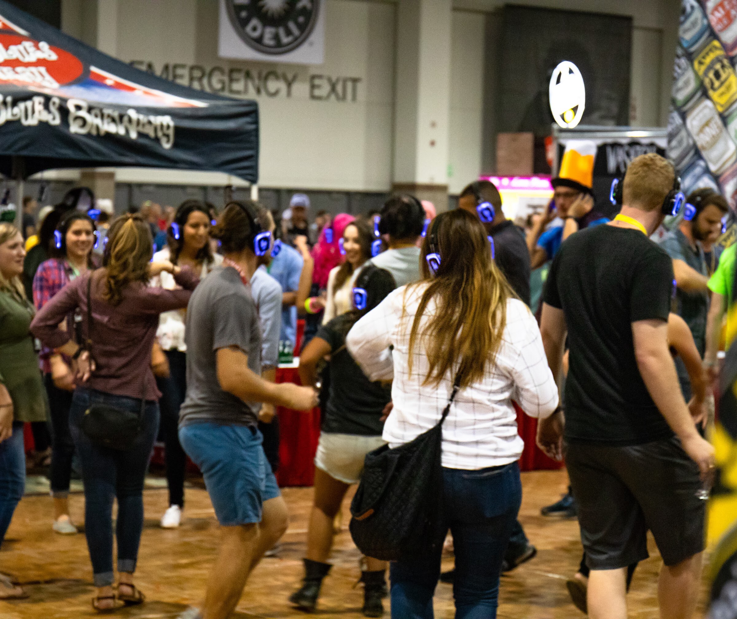 A group of people dancing on a wooden floor, several wearing headphones with blue lights, under an Emergency Exit sign and an Oskar Blues Brewery tent.
