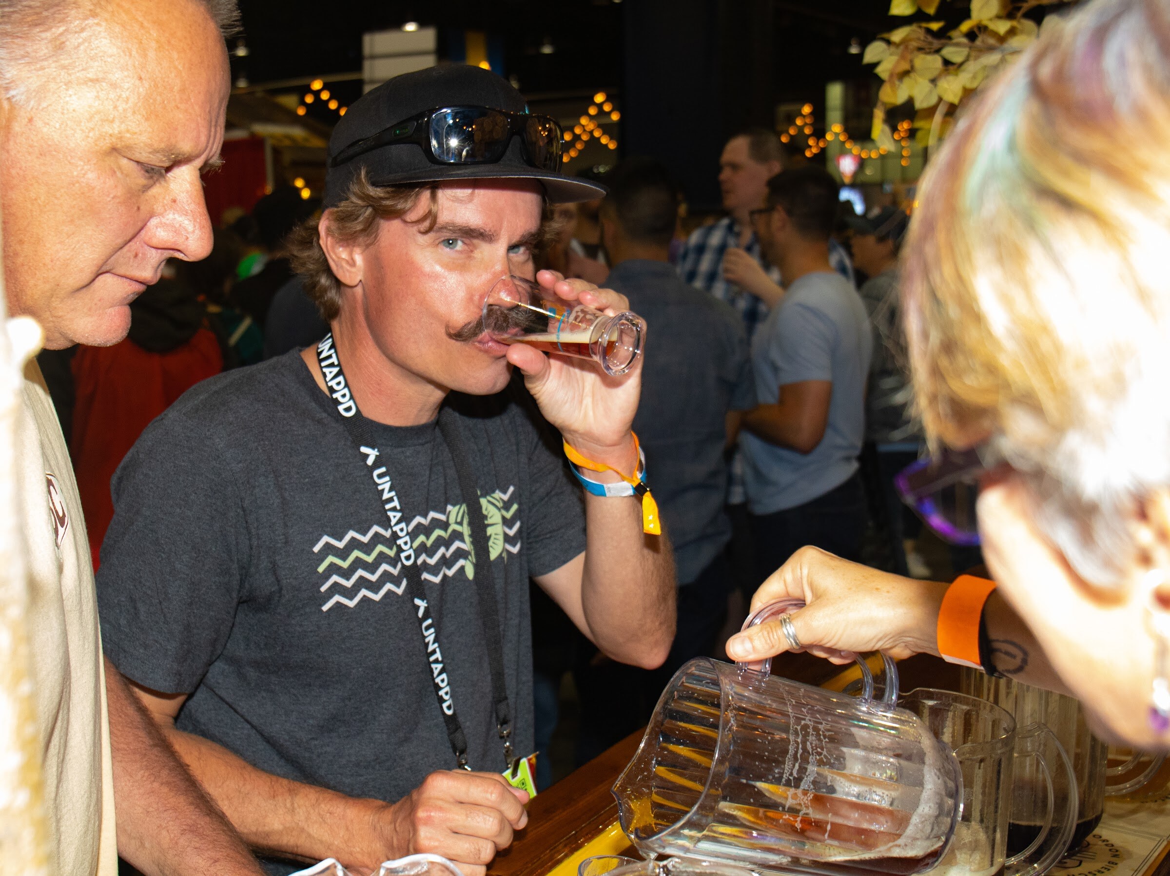 A man in a black cap with sunglasses and a gray T-shirt drinks from a small tasting glass at a counter while a pitcher of beer is being poured in the foreground.
