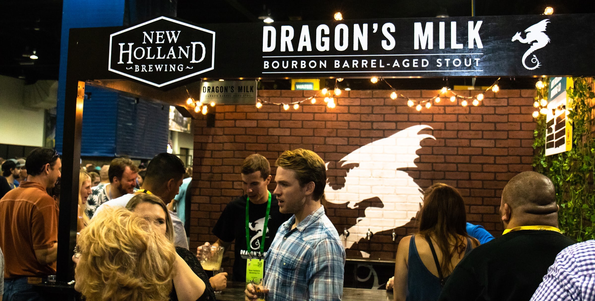 A black New Holland Brewing Dragon's Milk Bourbon Barrel-Aged Stout sign over a brick wall with a white dragon and string lights, with attendees and a server in front.