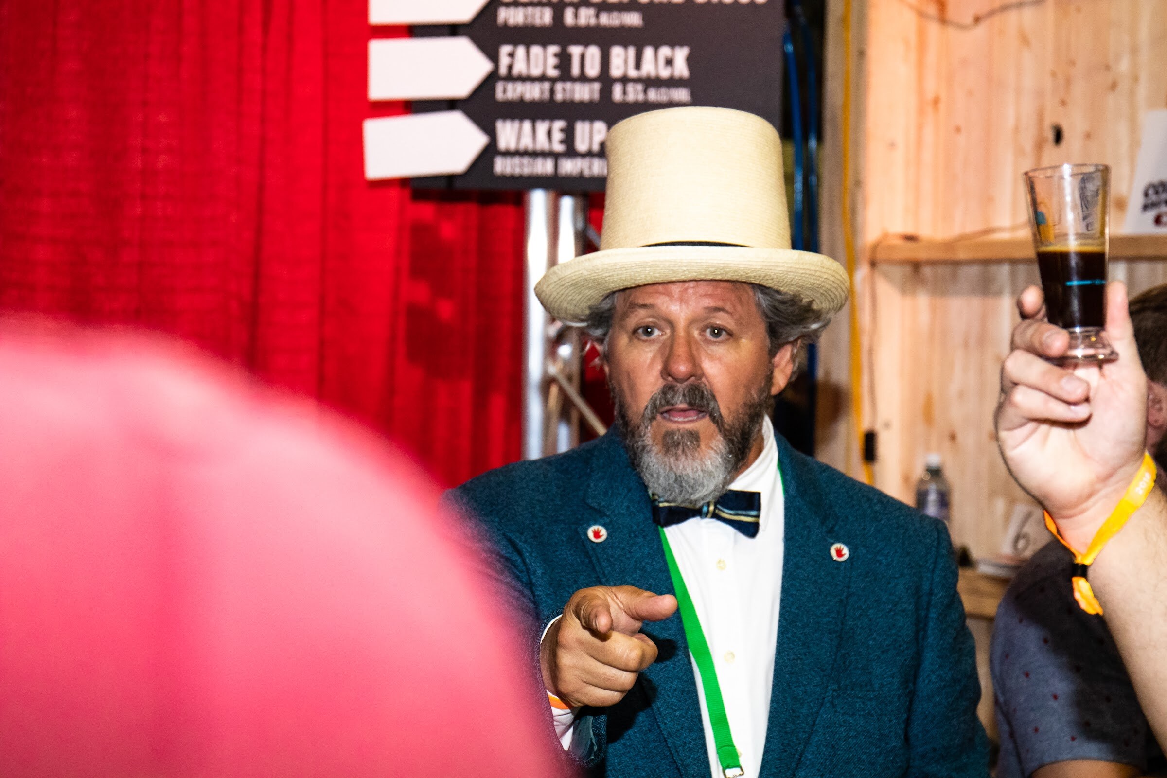 A bearded man in a teal blazer, bow tie and cream top hat points toward the camera beside a menu board listing Fade to Black export stout and Wake Up, with a hand holding up a tasting glass at right.