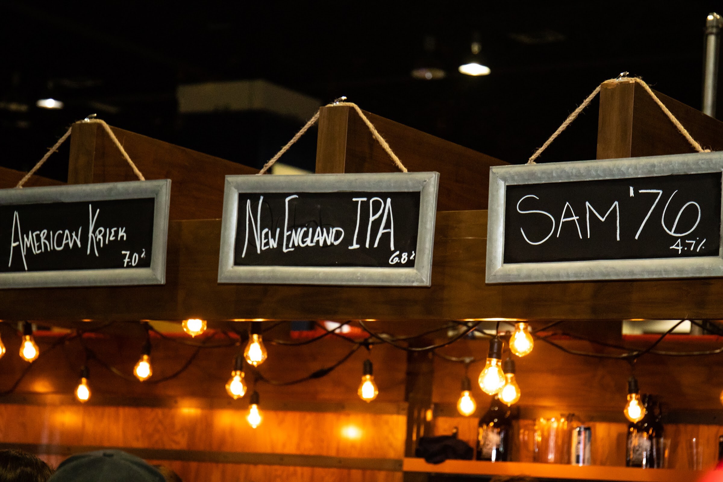 Three hanging chalkboard signs reading American Kriek, New England IPA and Sam '76 above a wooden bar lit by warm bulbs.